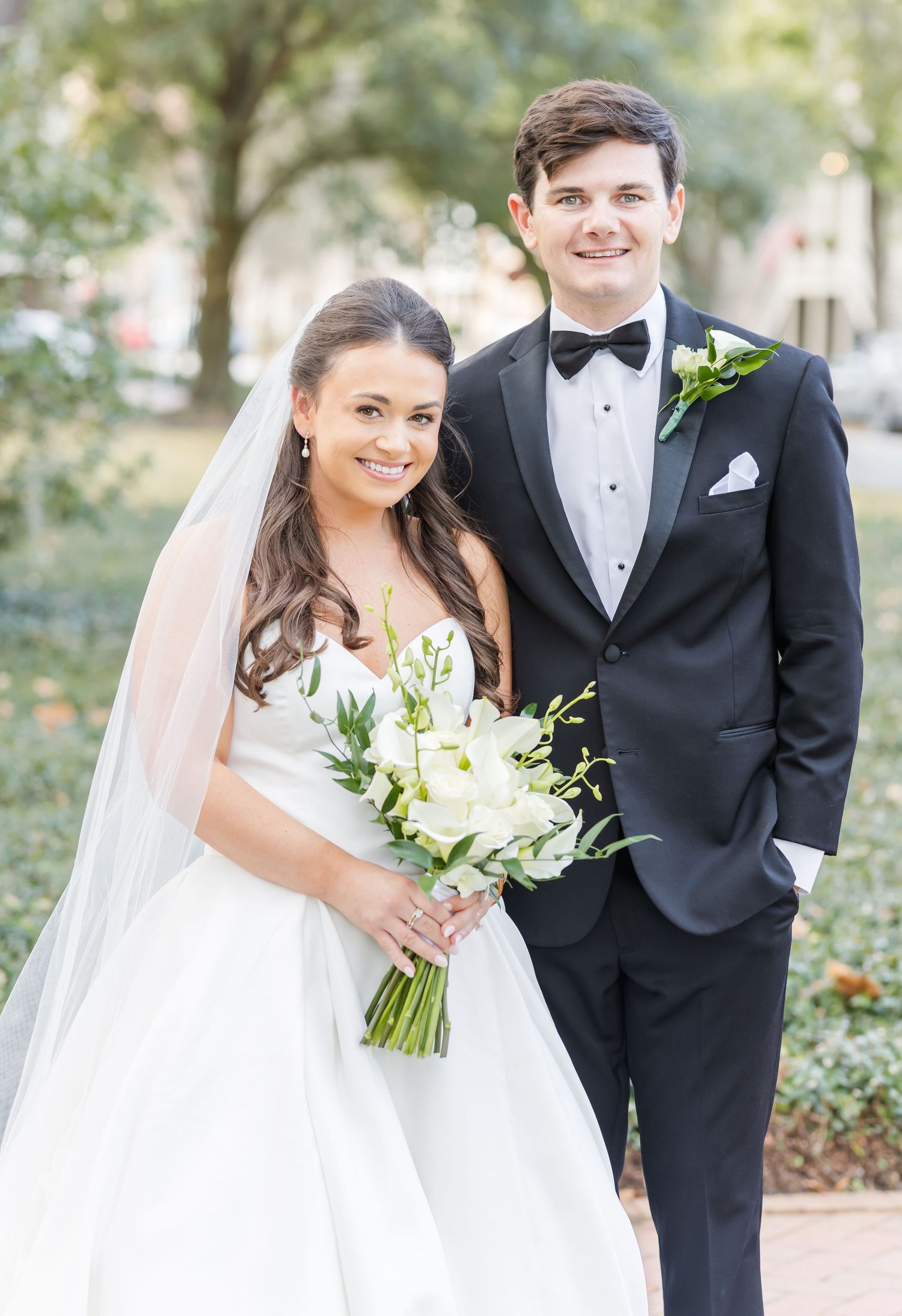 A bride and groom pose together outdoors in formal wedding attire with white floral bouquet and classic black tuxedo.