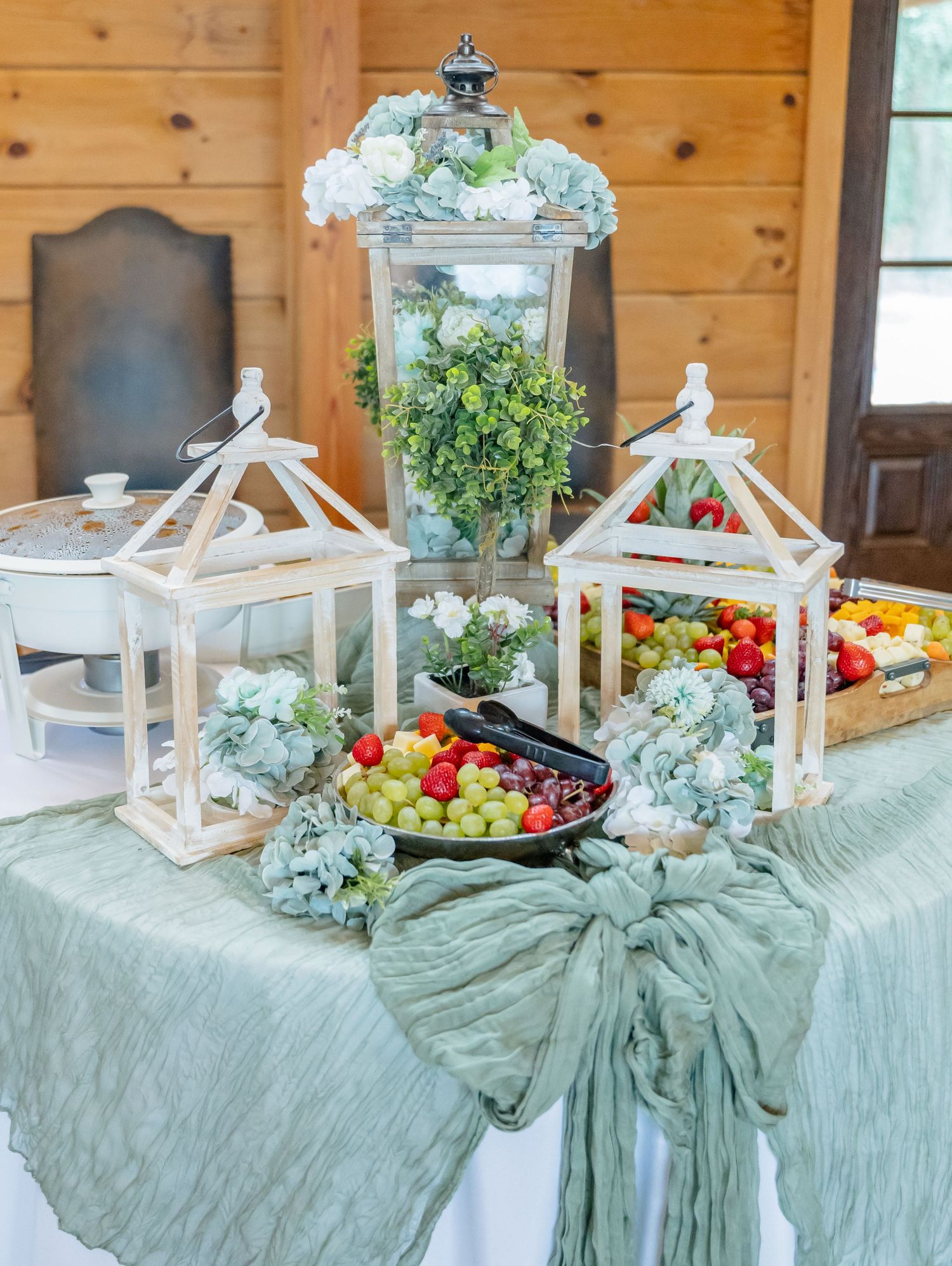 Elegant table display with white lanterns, fresh fruits, and sage green decor on a textured tablecloth for an event setting.