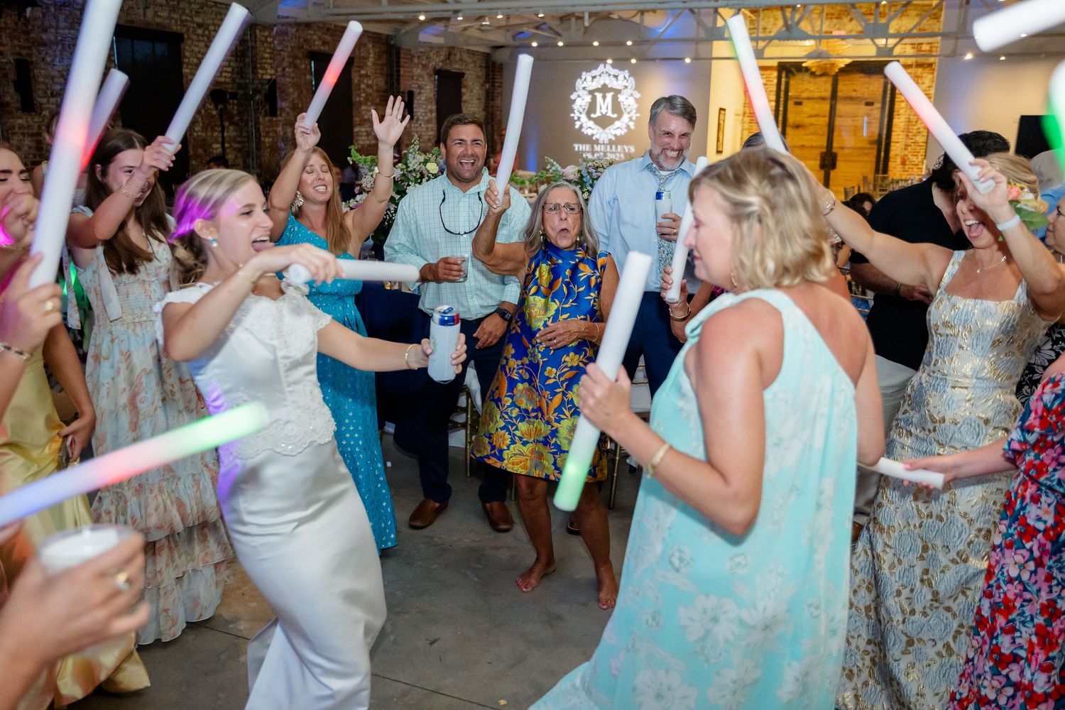 Wedding party dancing with glow sticks in a rustic brick venue with colorful lighting.
