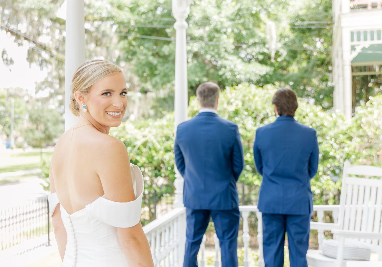 Wedding party in navy blue suits and white dress stand together on a porch in natural lighting.