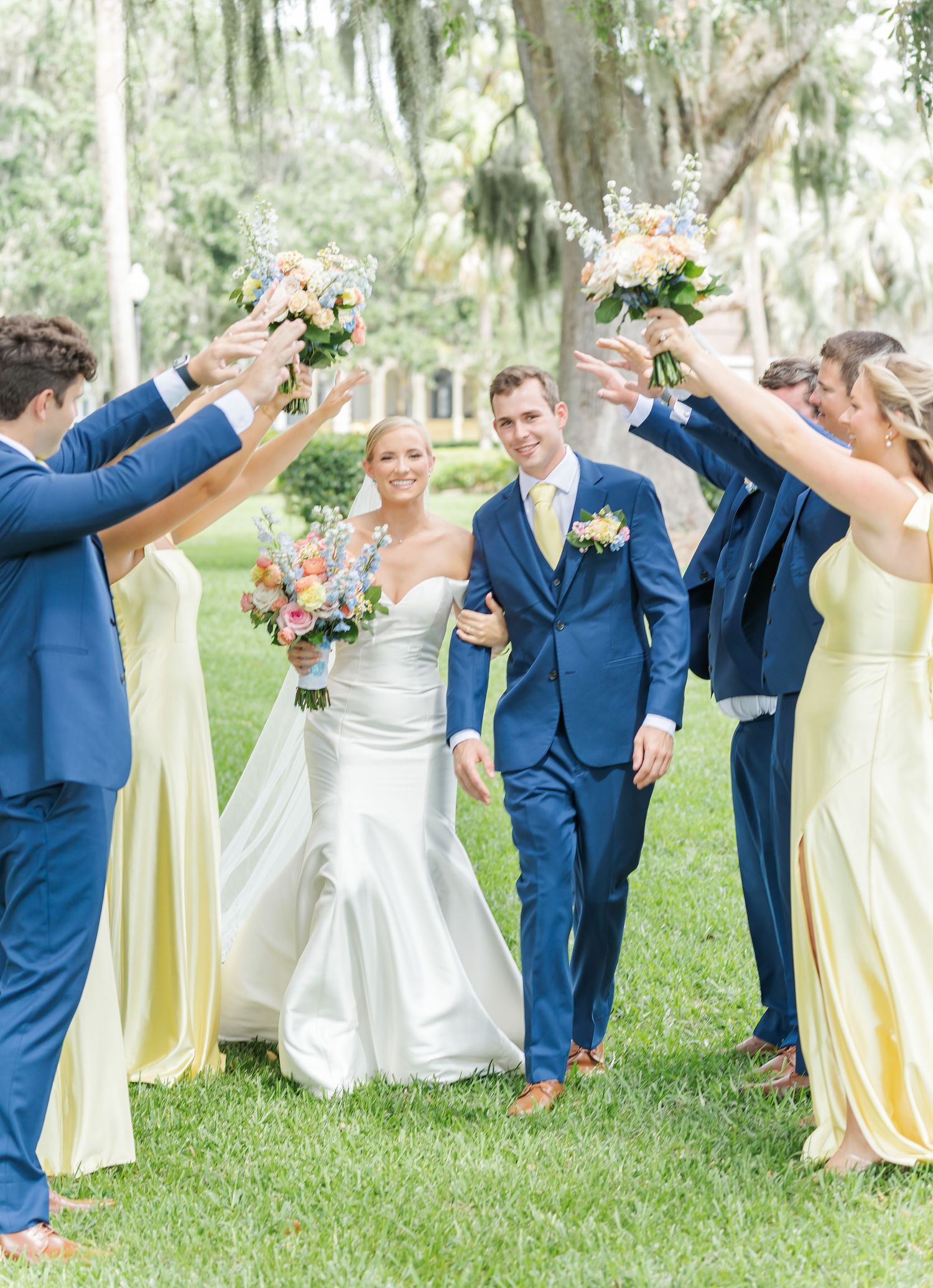 Wedding party in blue suits and yellow dresses poses playfully on grassy lawn under Spanish moss trees.