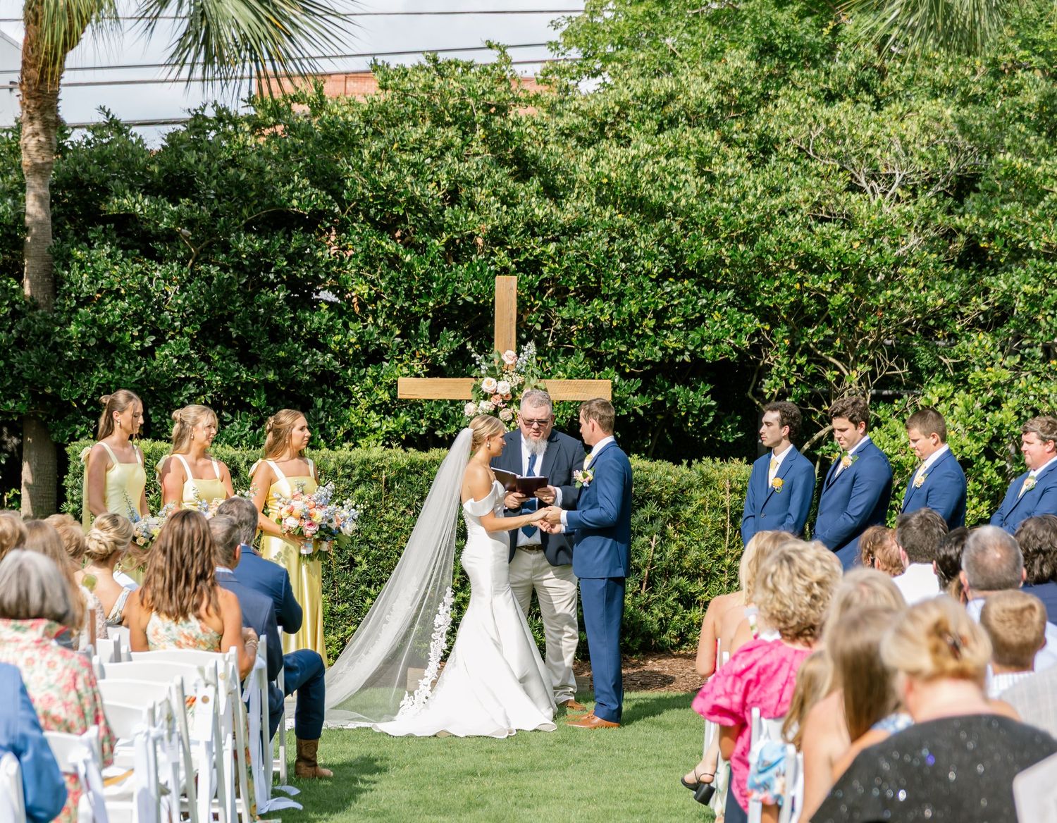 Outdoor wedding ceremony takes place in a lush garden setting with white chairs and tropical palm trees.