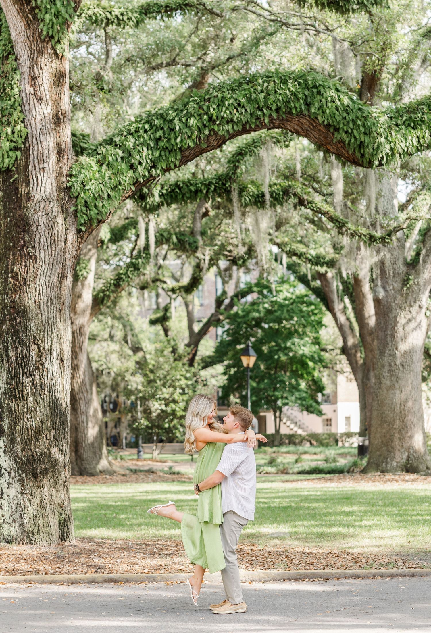 Couple embracing under moss-draped oak trees in a lush green park, woman in sage jumpsuit lifted off ground.