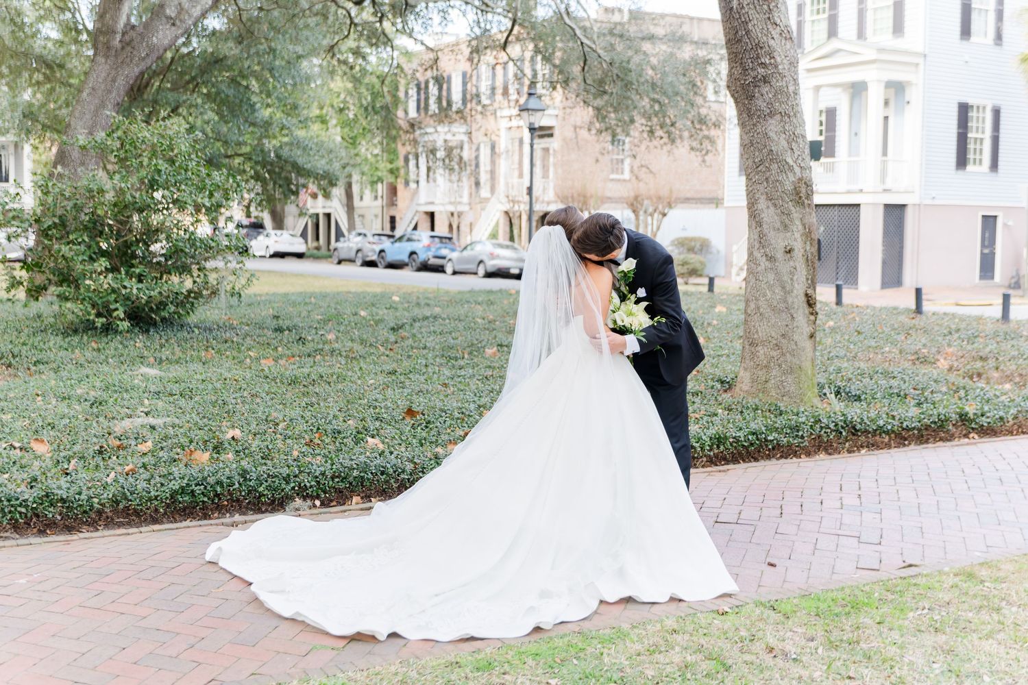 Bride and groom kiss on brick path, showcasing long cathedral wedding veil and bouquet.
