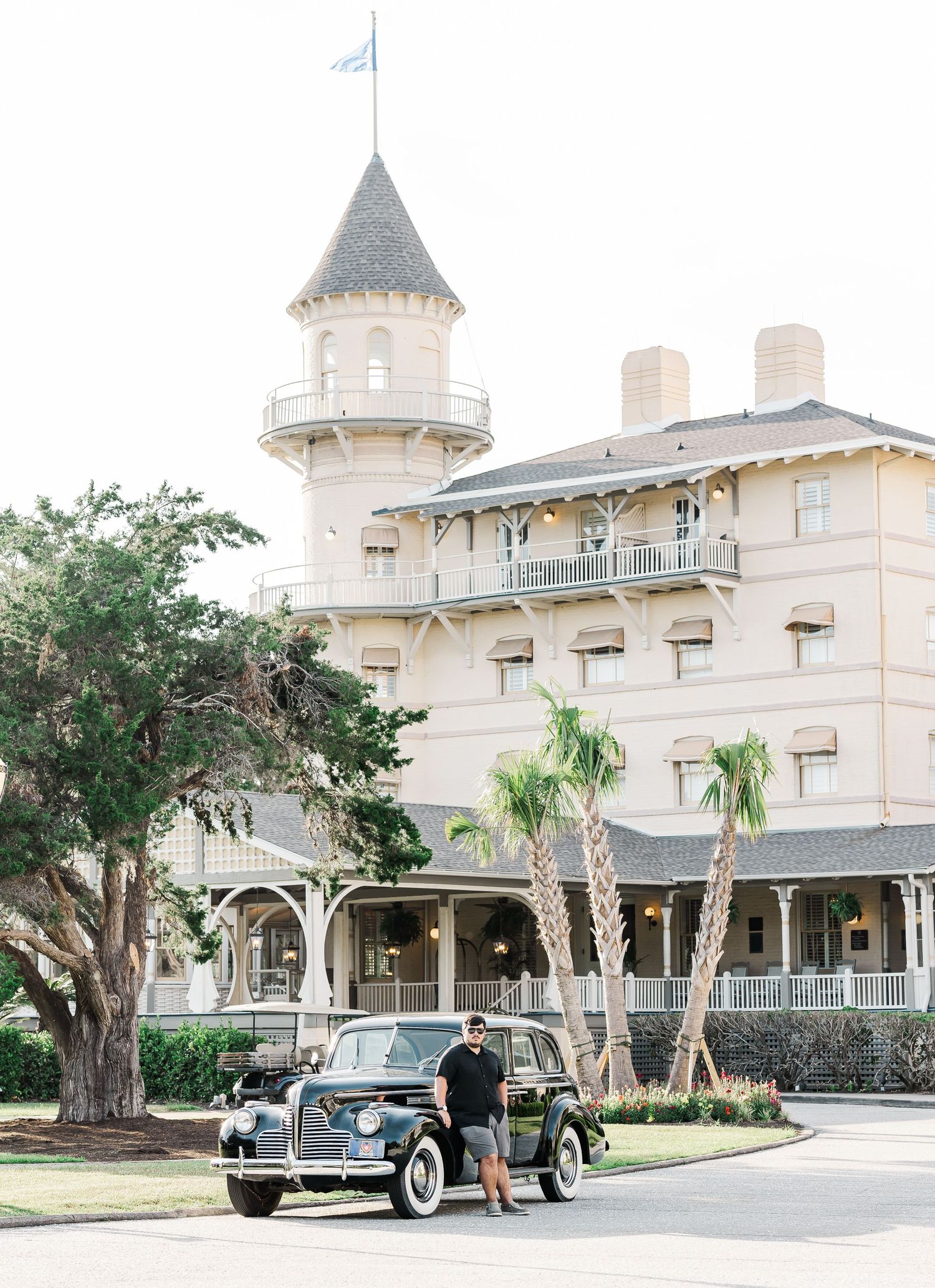 A vintage Rolls Royce luxury car parked in front of a Victorian-style mansion with turrets and palm trees.