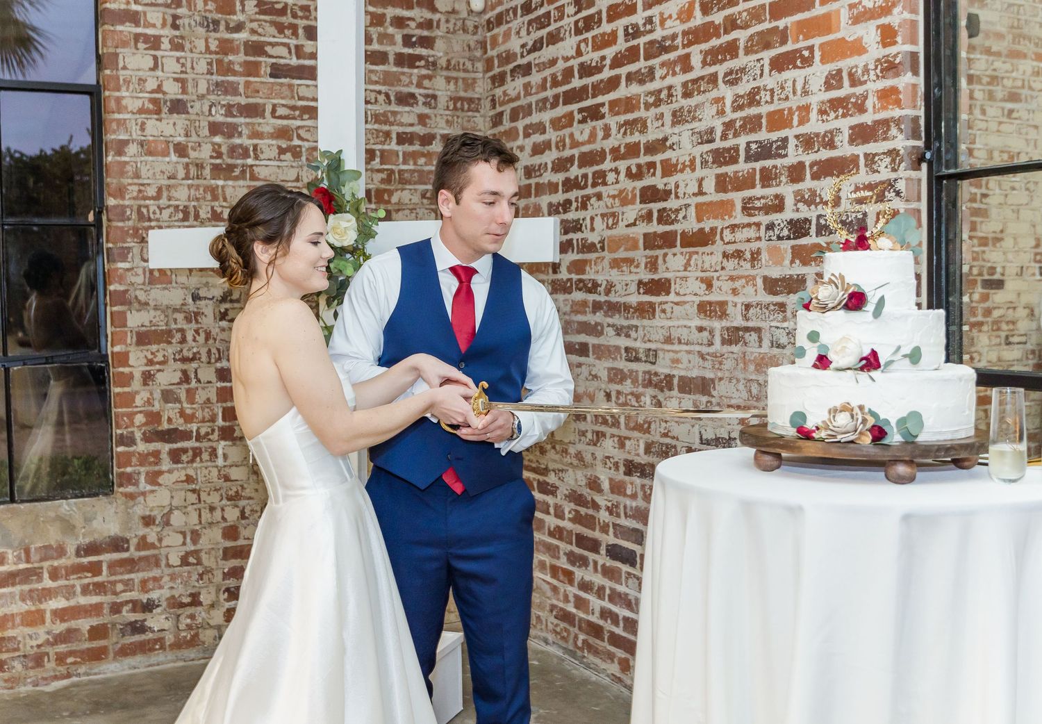 Couple in wedding attire cut their cake together at a rustic brick venue during their reception celebration.