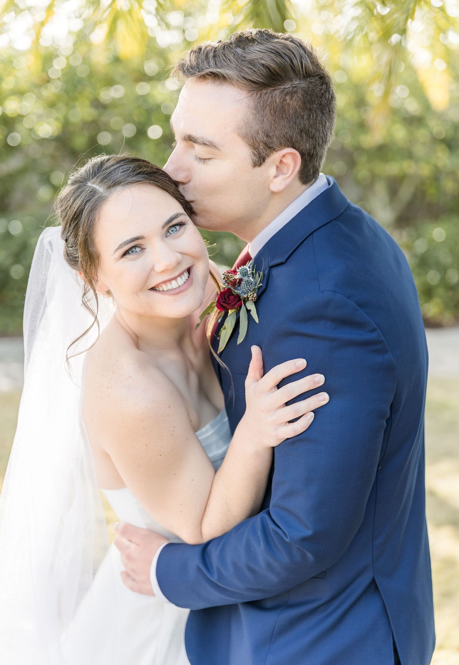 Wedding couple embraces outdoors in navy suit and white dress with natural lighting.