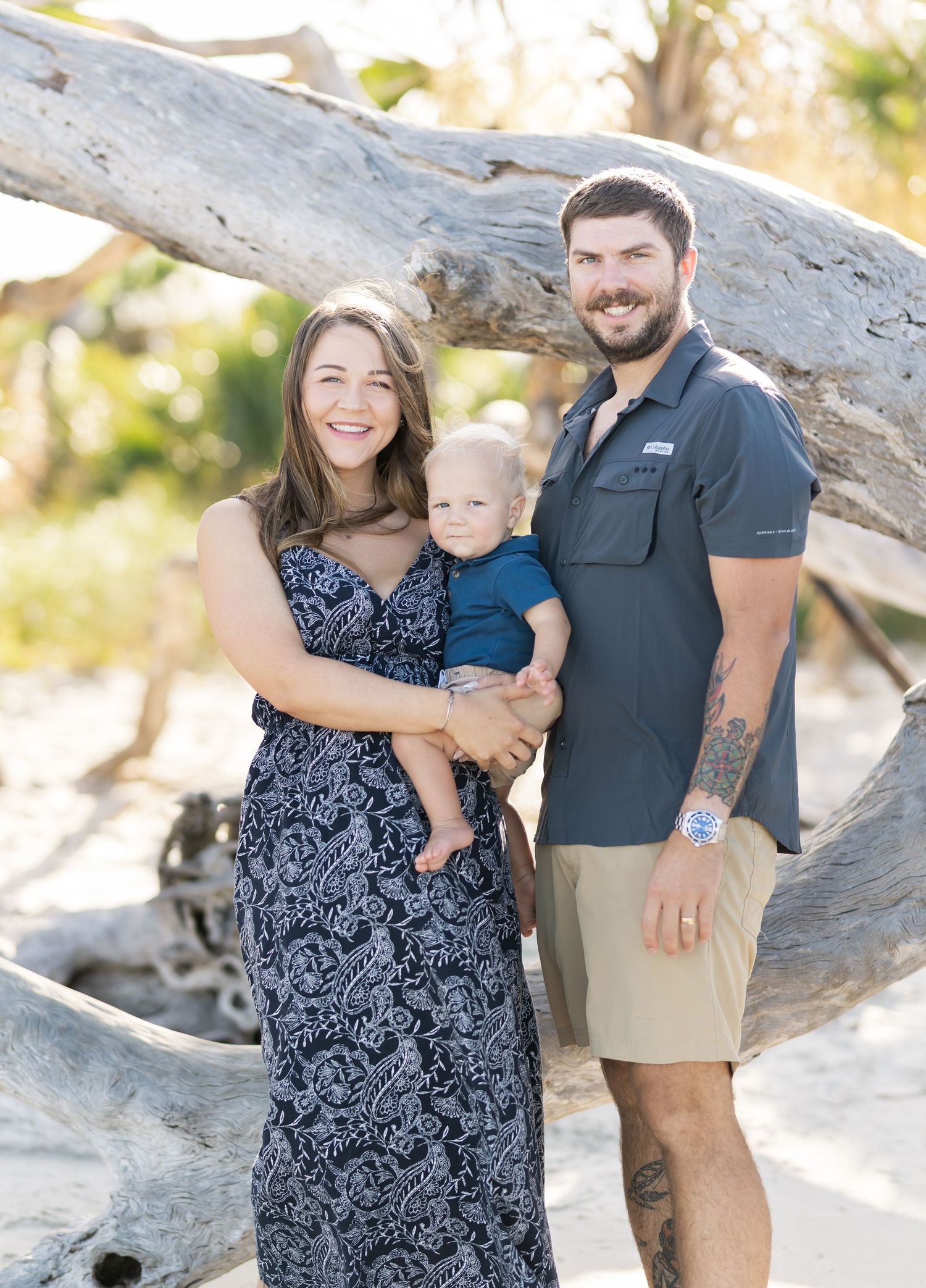 A family poses together for a portrait against a driftwood log backdrop on a sunny beach.