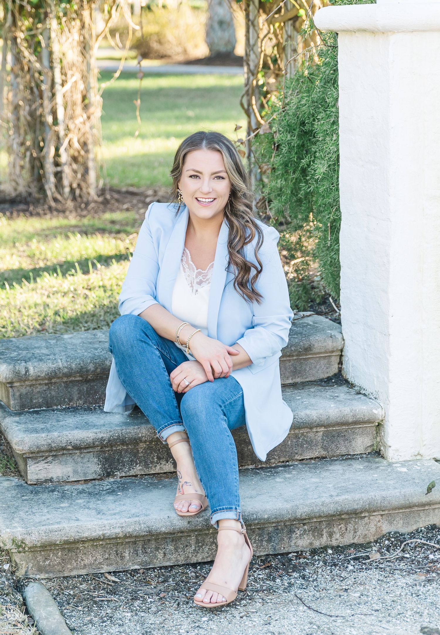 A person wearing a light blue blazer and jeans sits casually on stone steps outdoors on a sunny day.