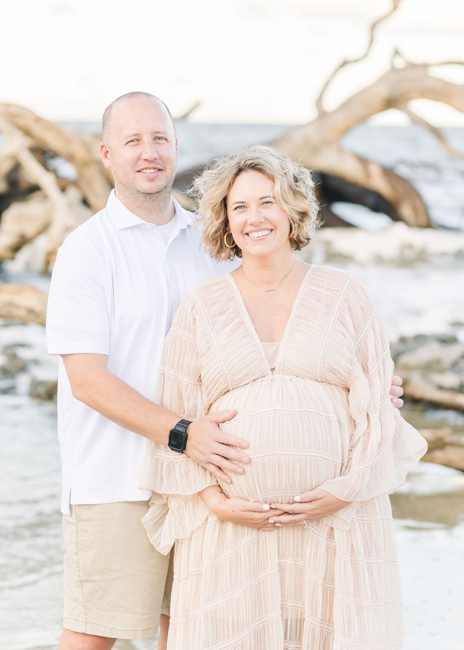 Expectant couple shares tender moment on beach with driftwood and ocean in background.