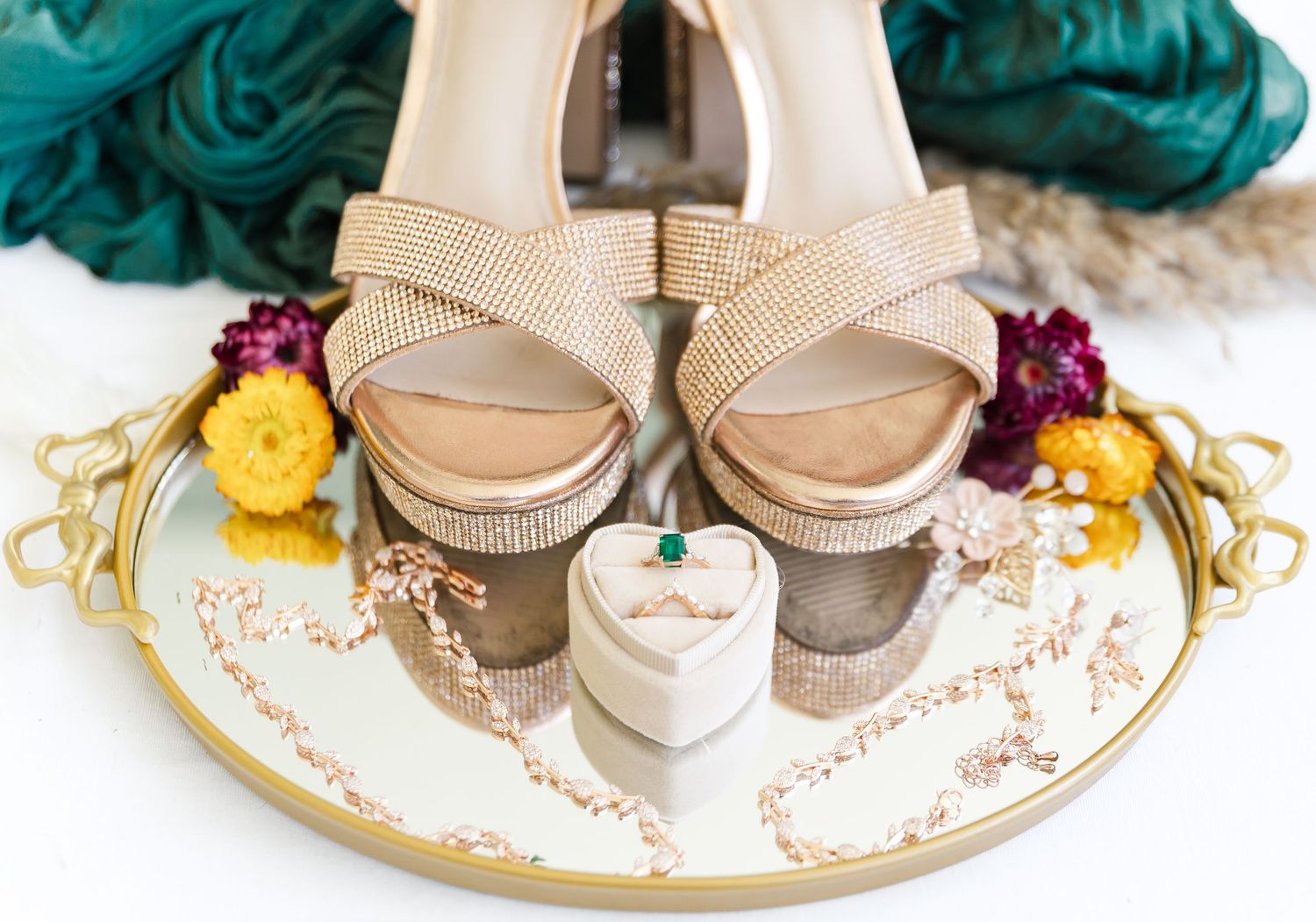 Sparkling gold wedding shoes displayed on a decorative mirrored tray with colorful flowers and ring box.