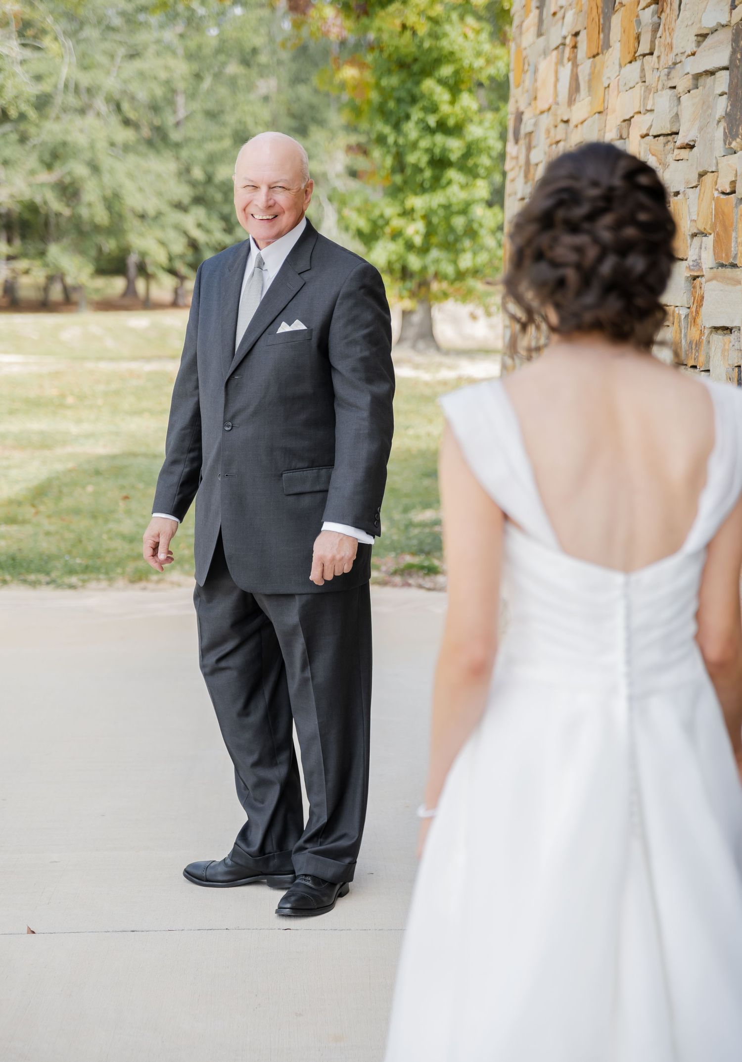 A first look moment between a father and bride in formal attire standing by a stone pillar outdoors.