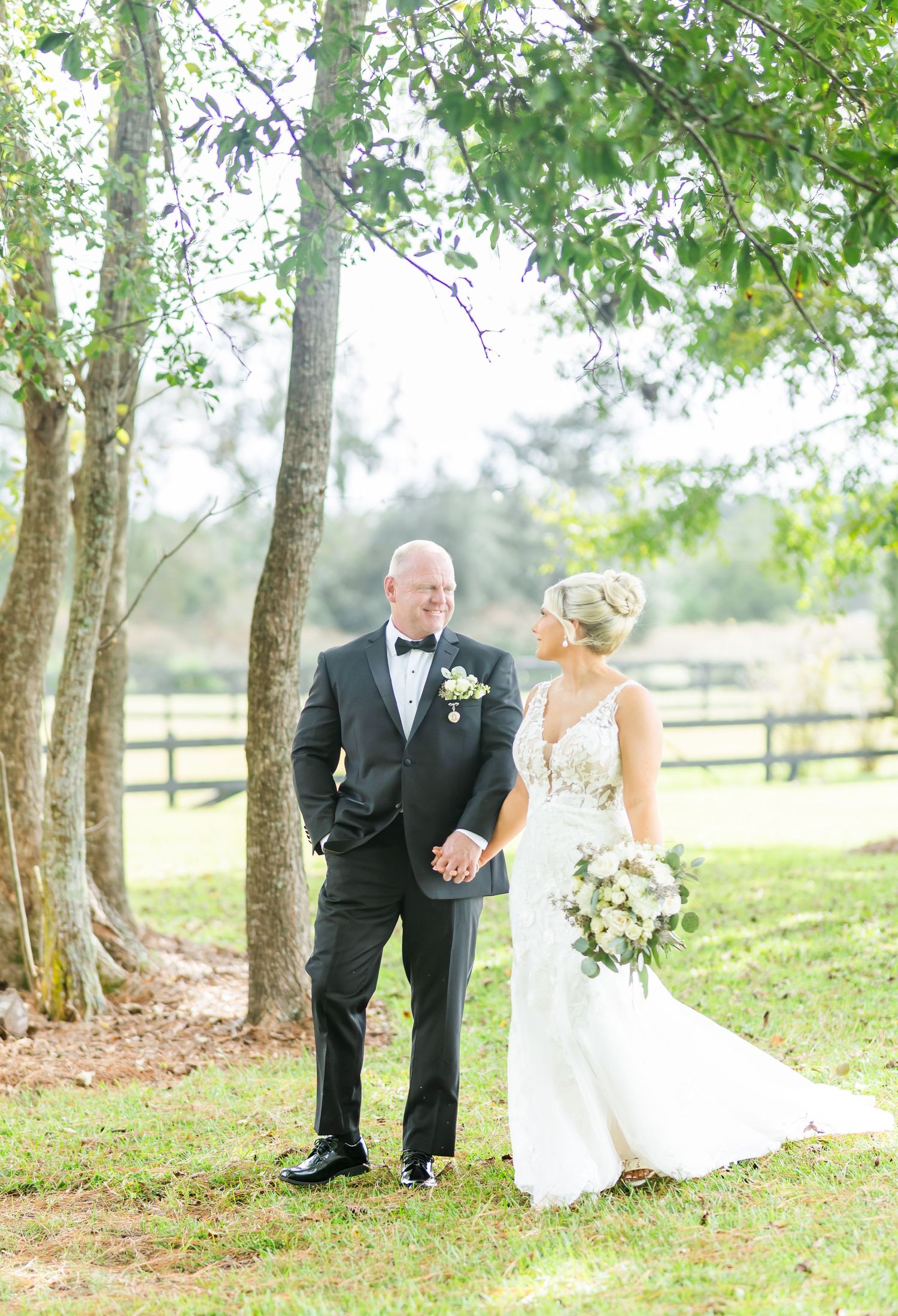 A couple poses together outdoors under trees on their wedding day, with a rustic fence and greenery in the background.