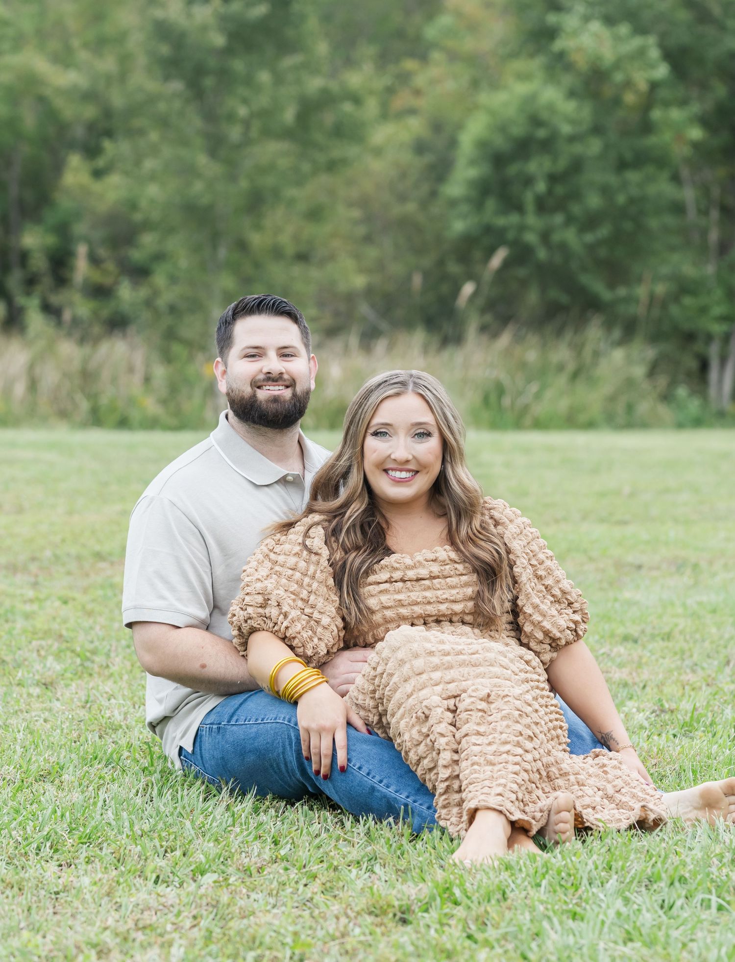 A couple lounges together on green grass during an outdoor photo session wearing casual attire and warm earth tones.