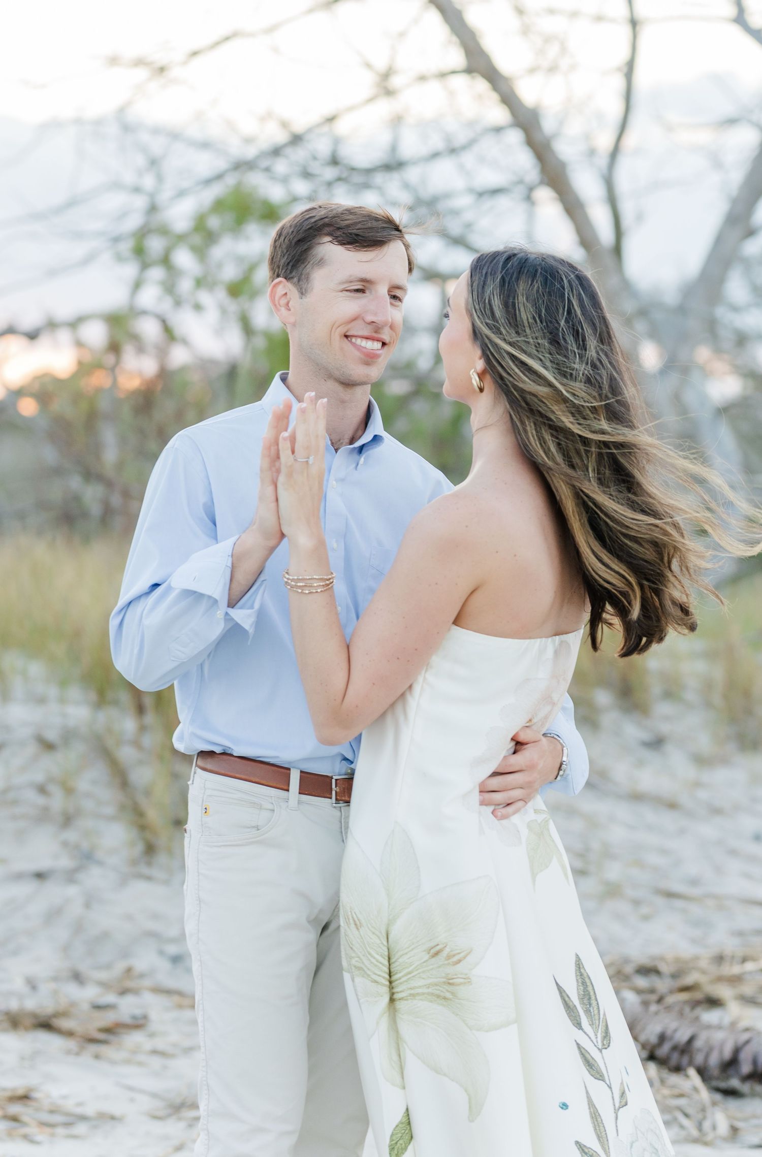 Couple dancing and smiling on sandy beach at dusk with windswept hair and soft lighting.