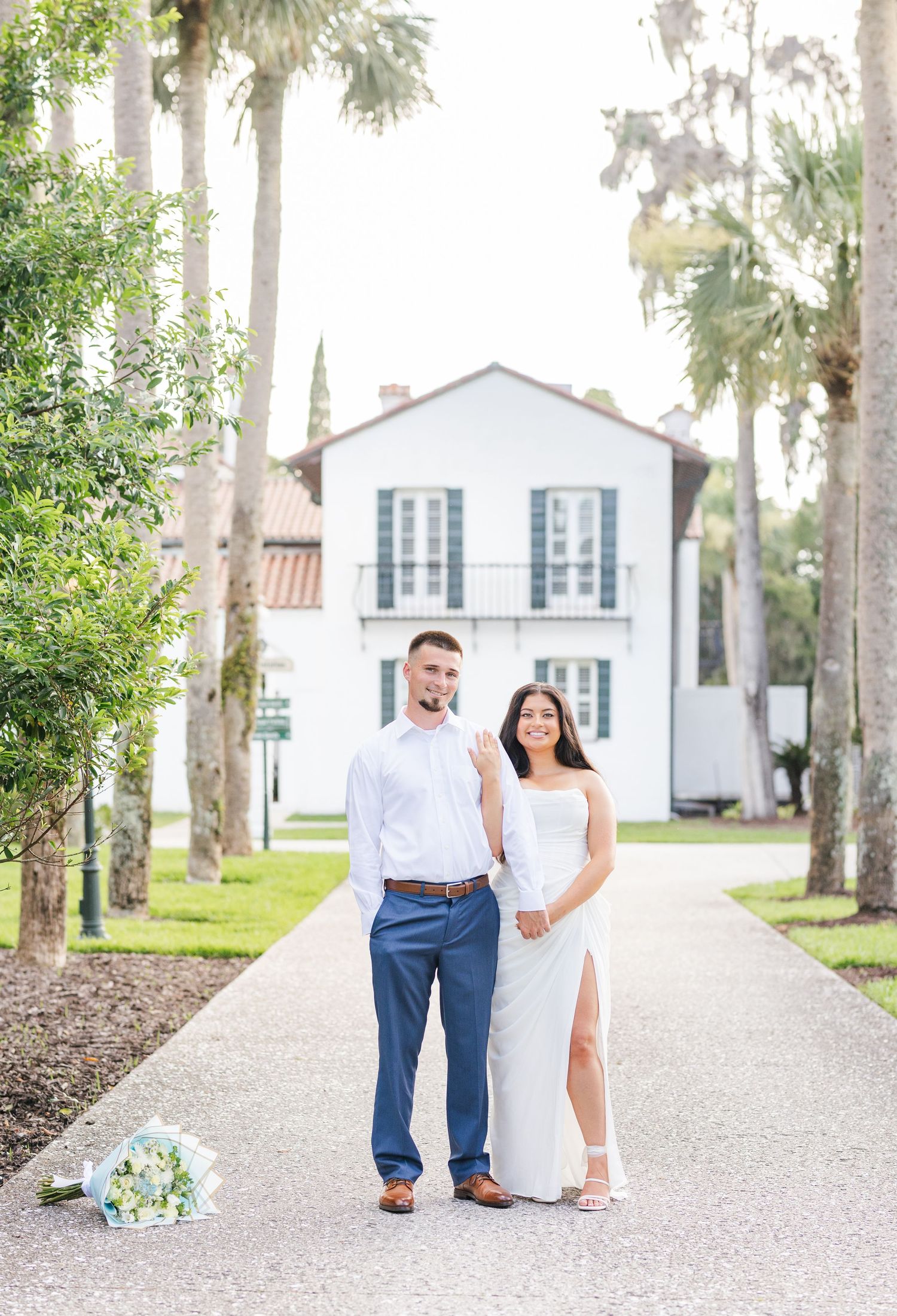 Engaged couple in white dress and blue pants pose on tree-lined path before historic white building.
