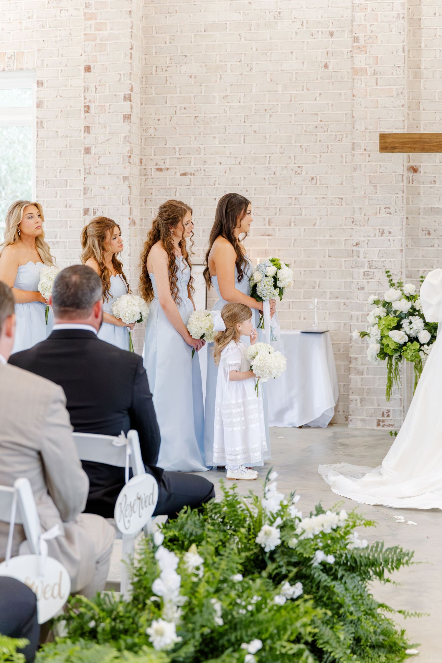 Bridesmaids in light blue gowns holding white bouquets stand at the altar with a small flower girl.