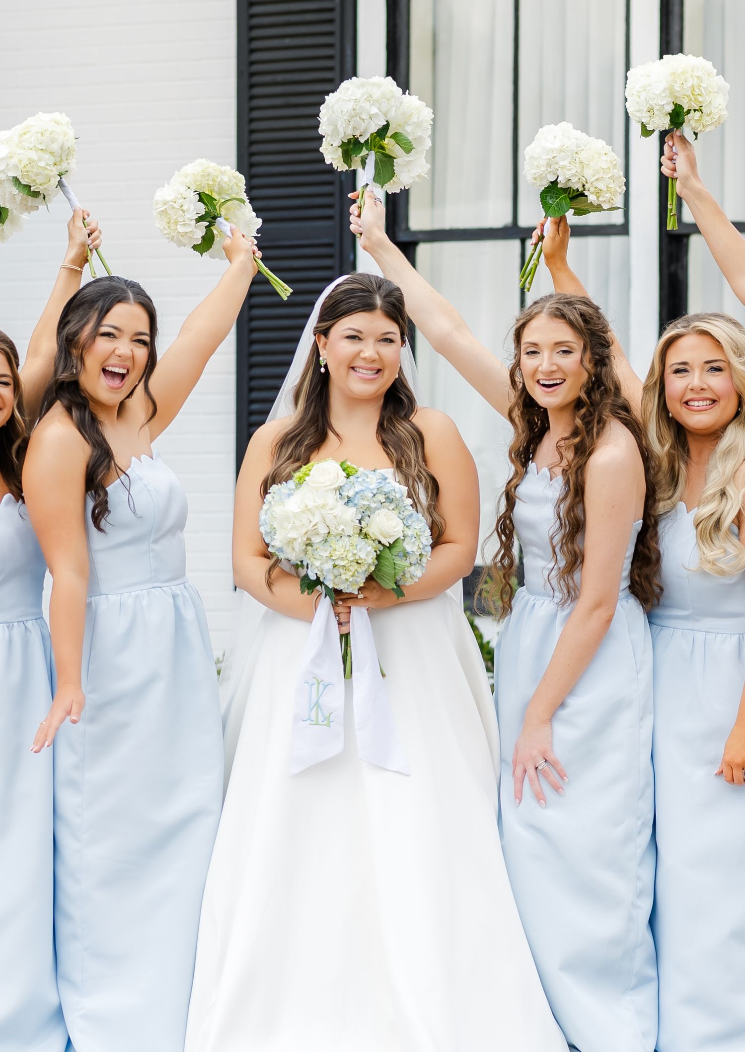 Joyful bride and bridesmaids in light blue dresses raise white hydrangea bouquets in celebration.