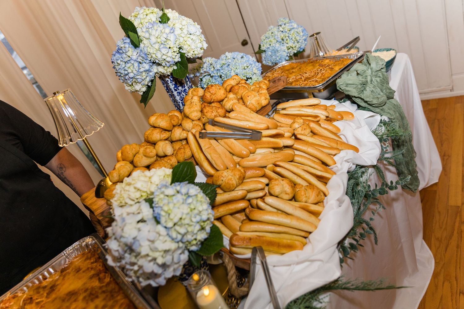 Elegant wedding reception bread and breadstick display arranged on a white tablecloth with blue hydrangea floral arrangements.