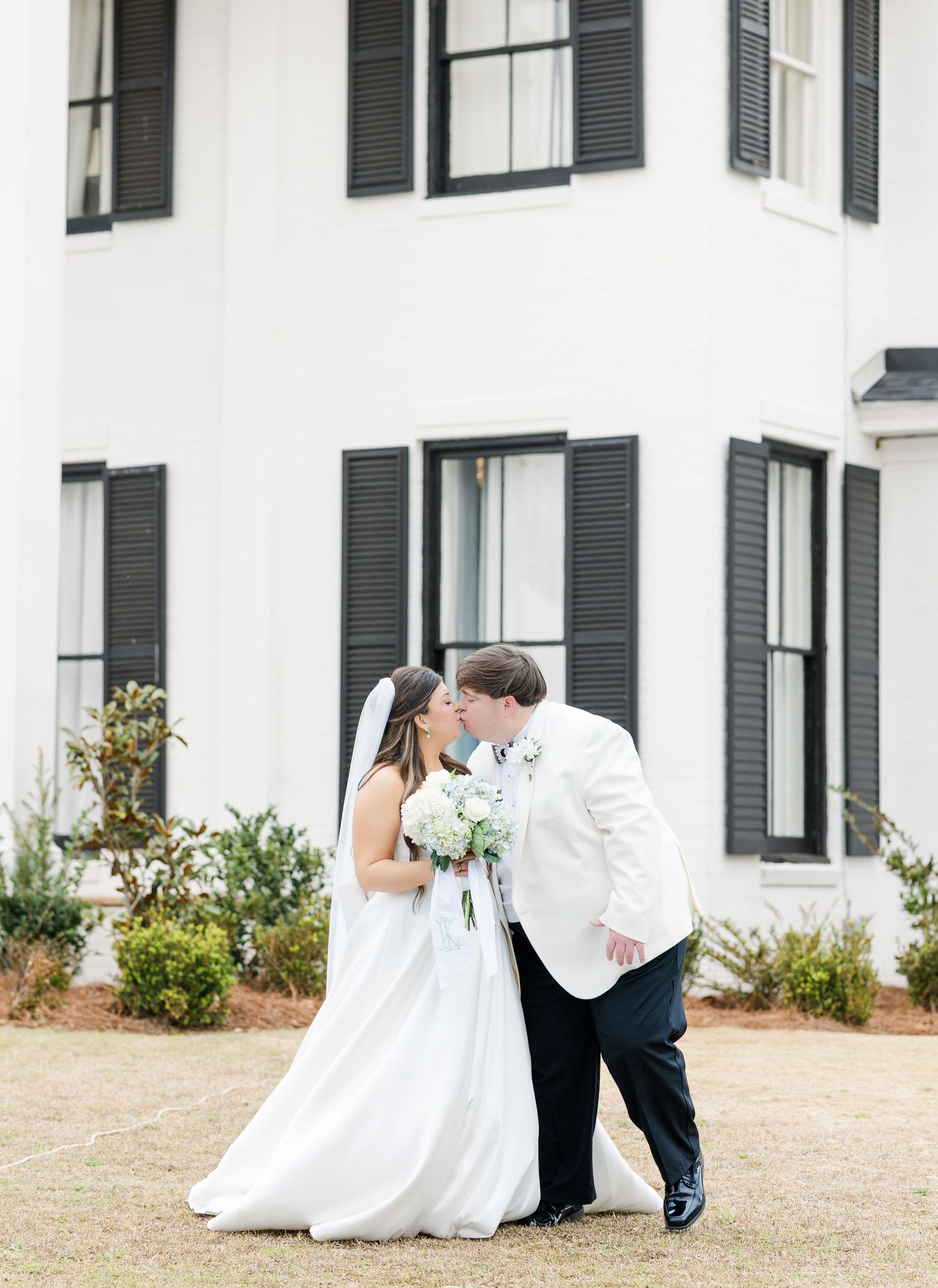Bride and groom share a kiss outside elegant white Southern mansion with classic black shutters and manicured grounds.