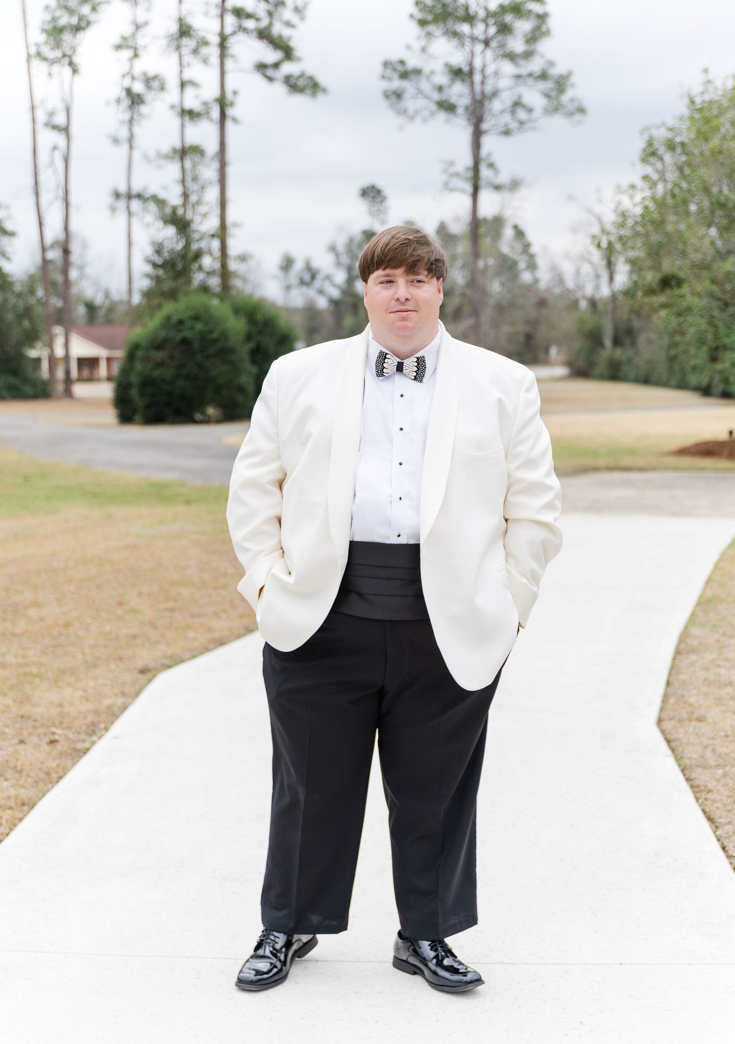Groom in white tuxedo jacket, black pants, and bow tie standing on a pathway outdoors.