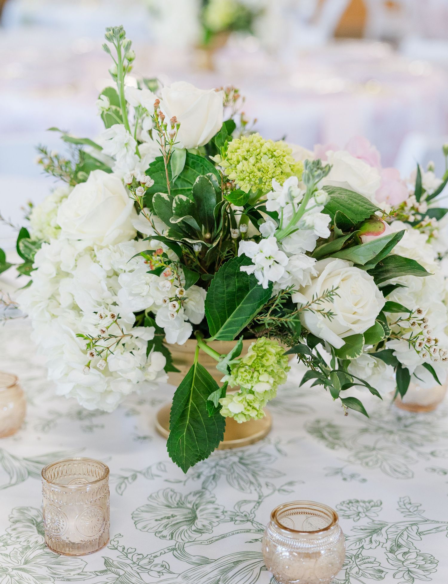 White roses and hydrangeas with green foliage in a gold vase centerpiece on a floral tablecloth at a wedding reception.