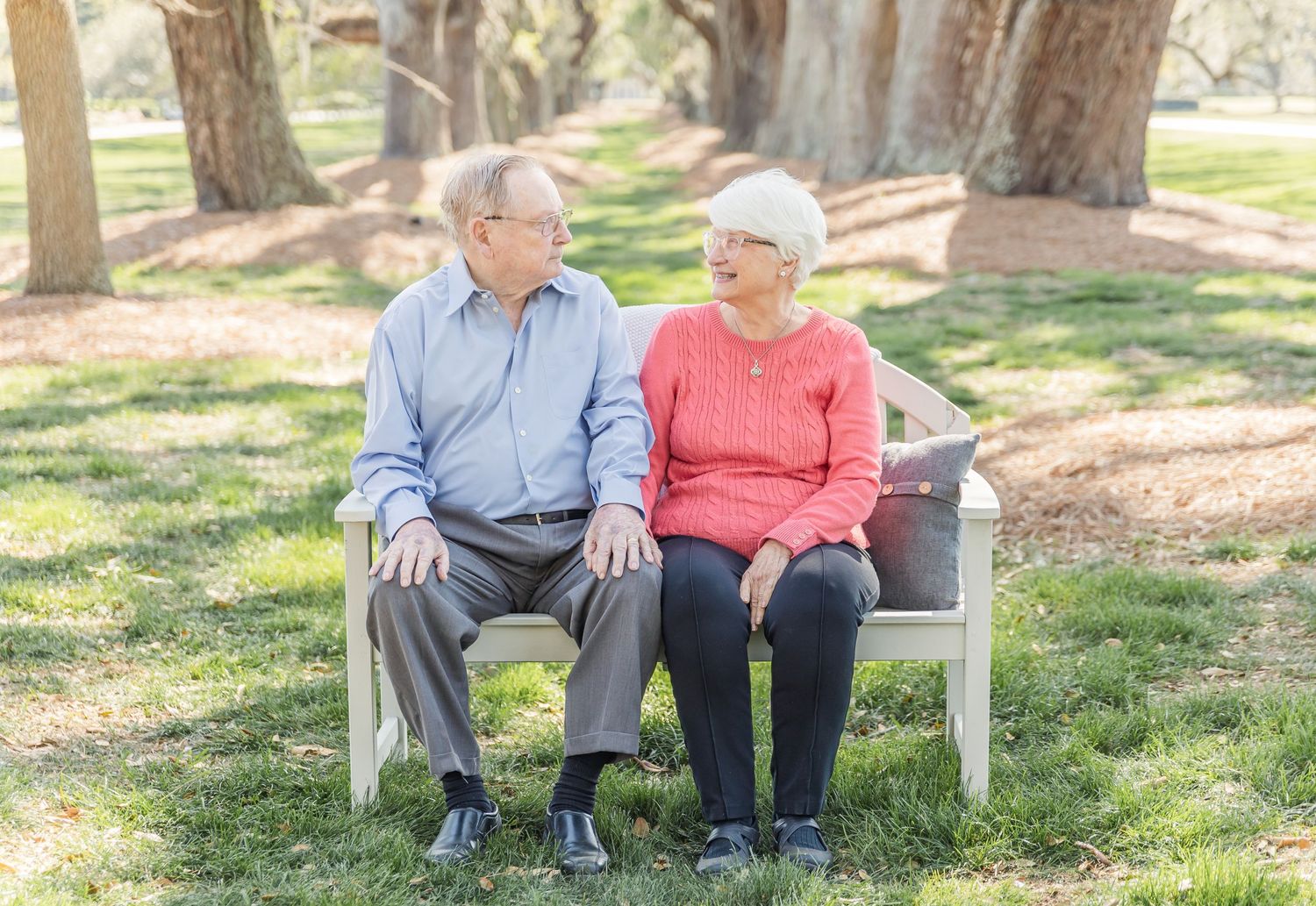 Elderly couple sits together on a white bench in a sunlit park setting.