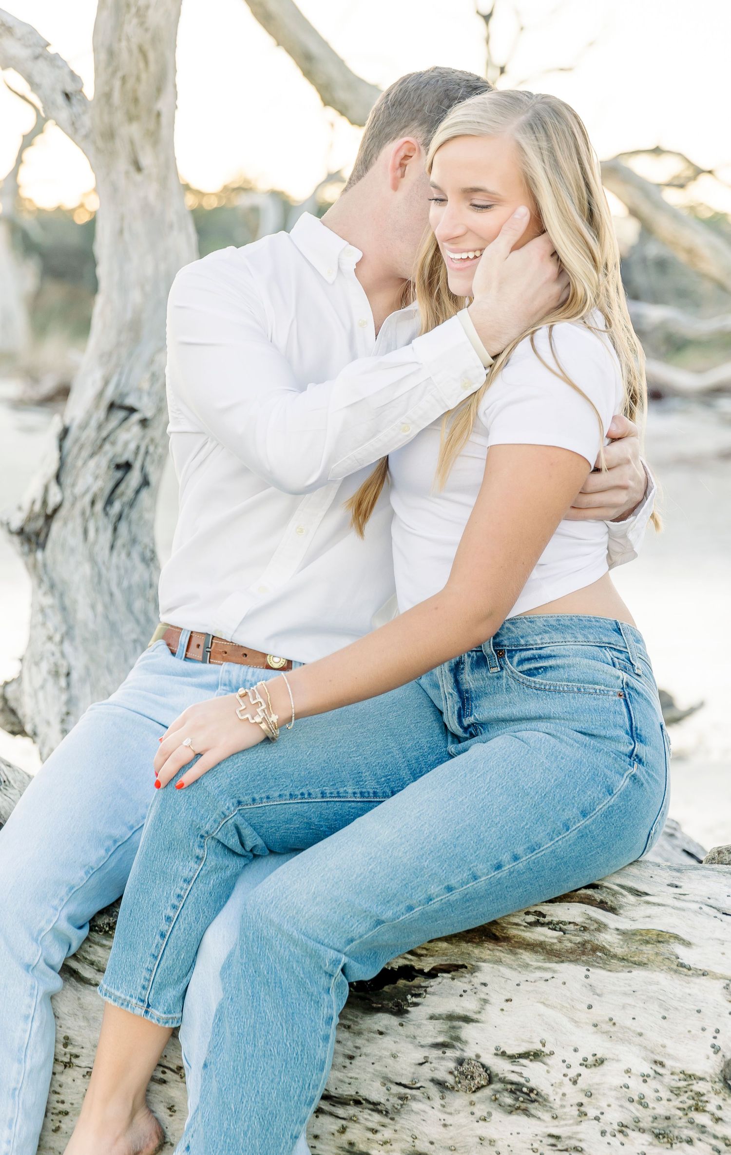 Romantic beach photo of couple in white and denim sharing a tender moment on driftwood.