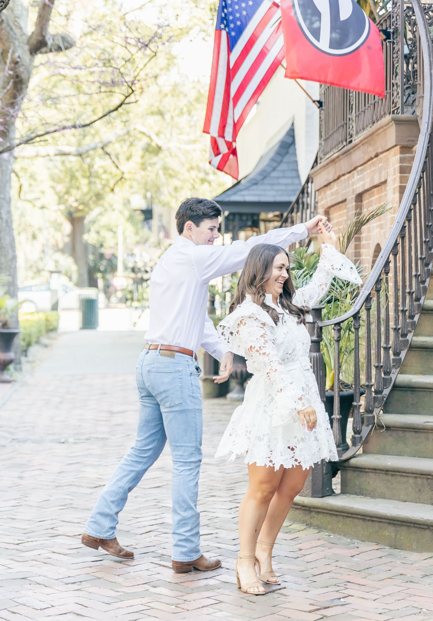 A couple poses playfully on brick steps with American flag and state flag hanging overhead.