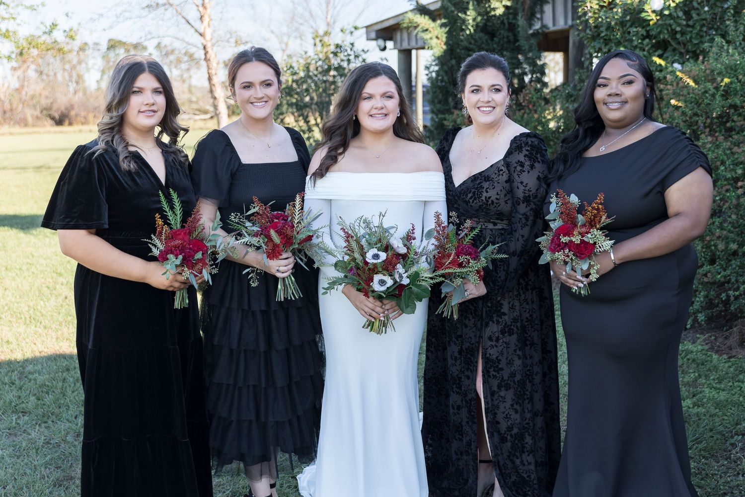 Bridesmaids in black gowns stand with bride in white dress holding red and white floral bouquets.