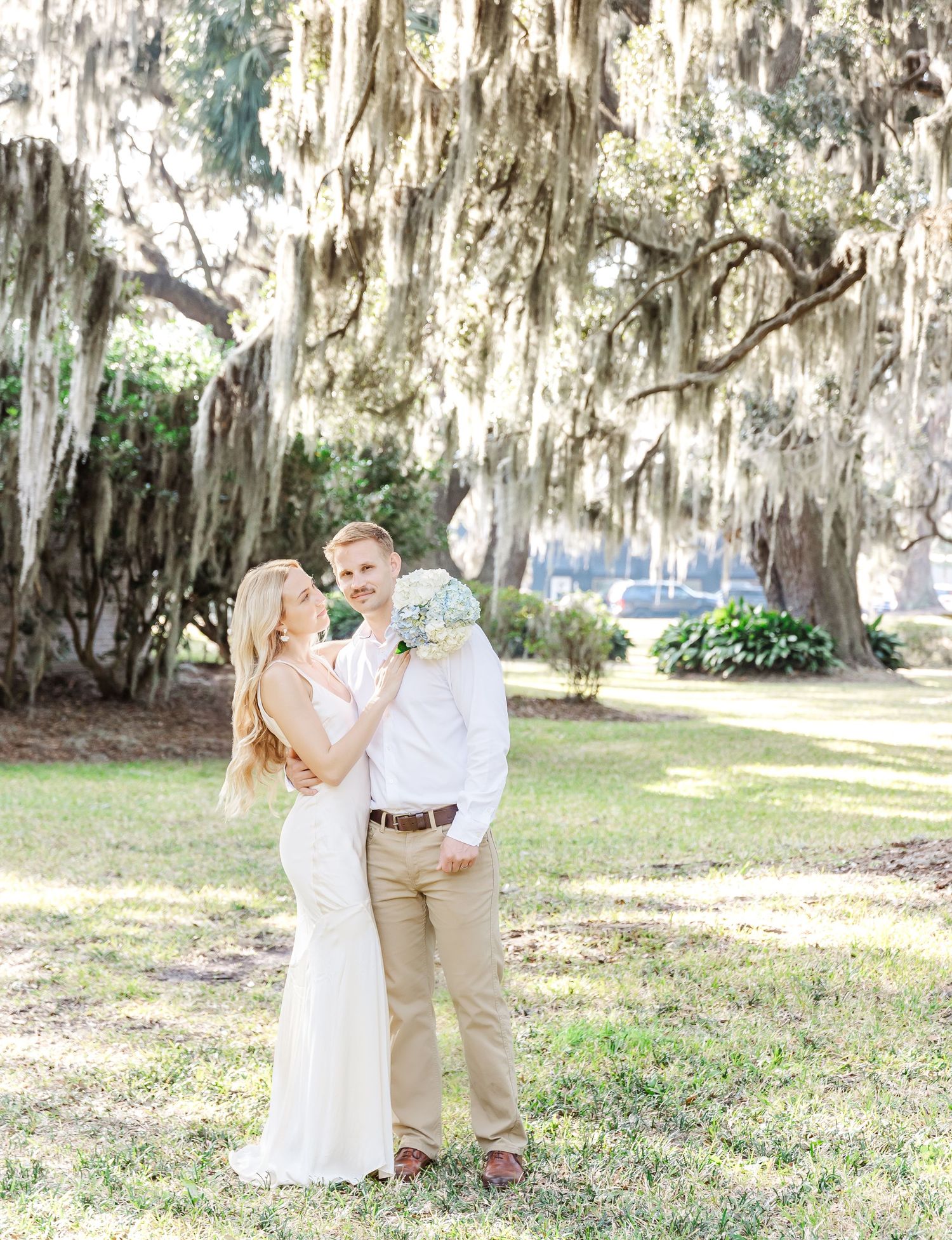Wedding couple shares an intimate moment under moss-covered trees in a sunlit garden setting.