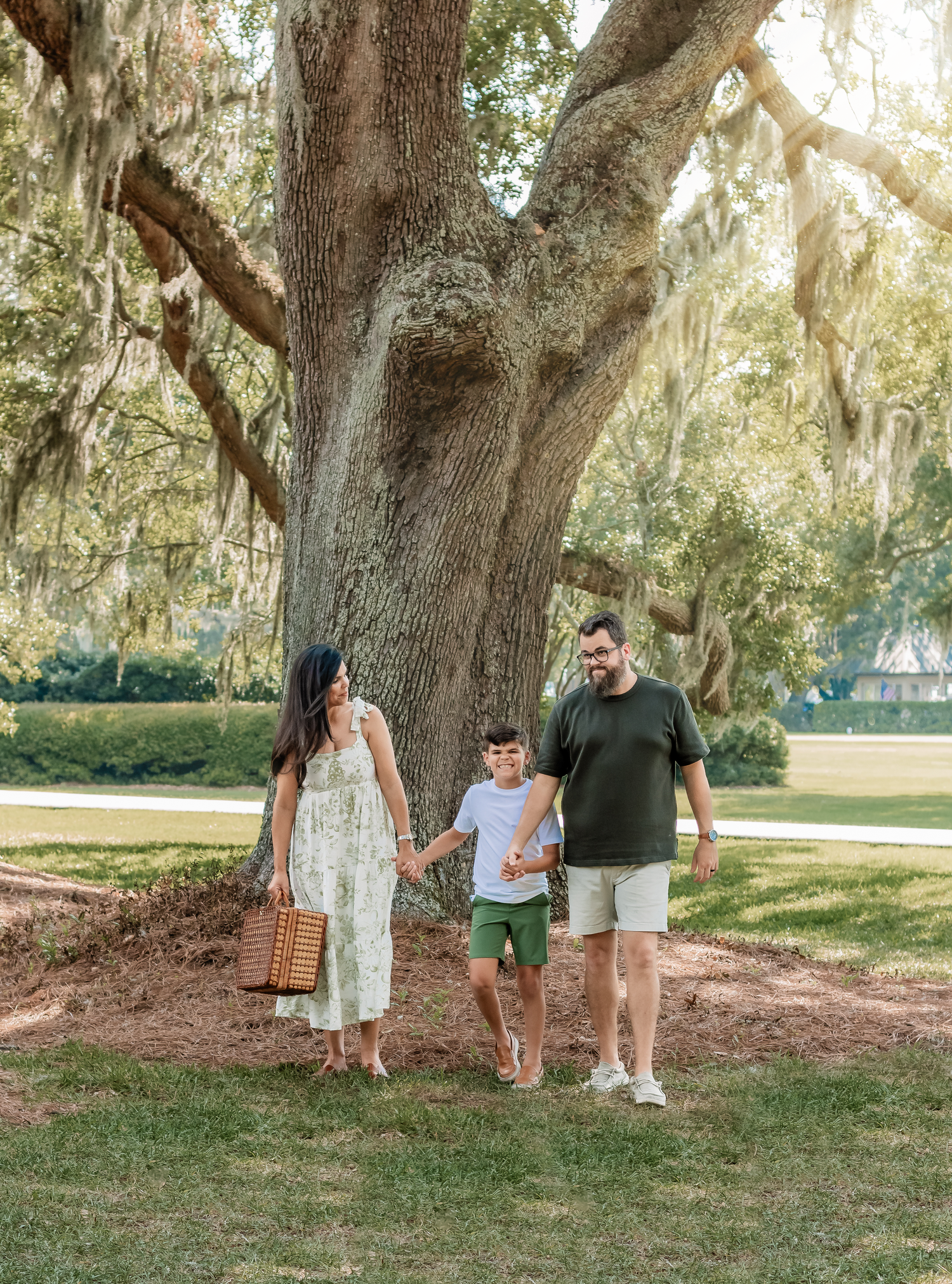 Family walks together under large oak trees with Spanish moss in park setting.