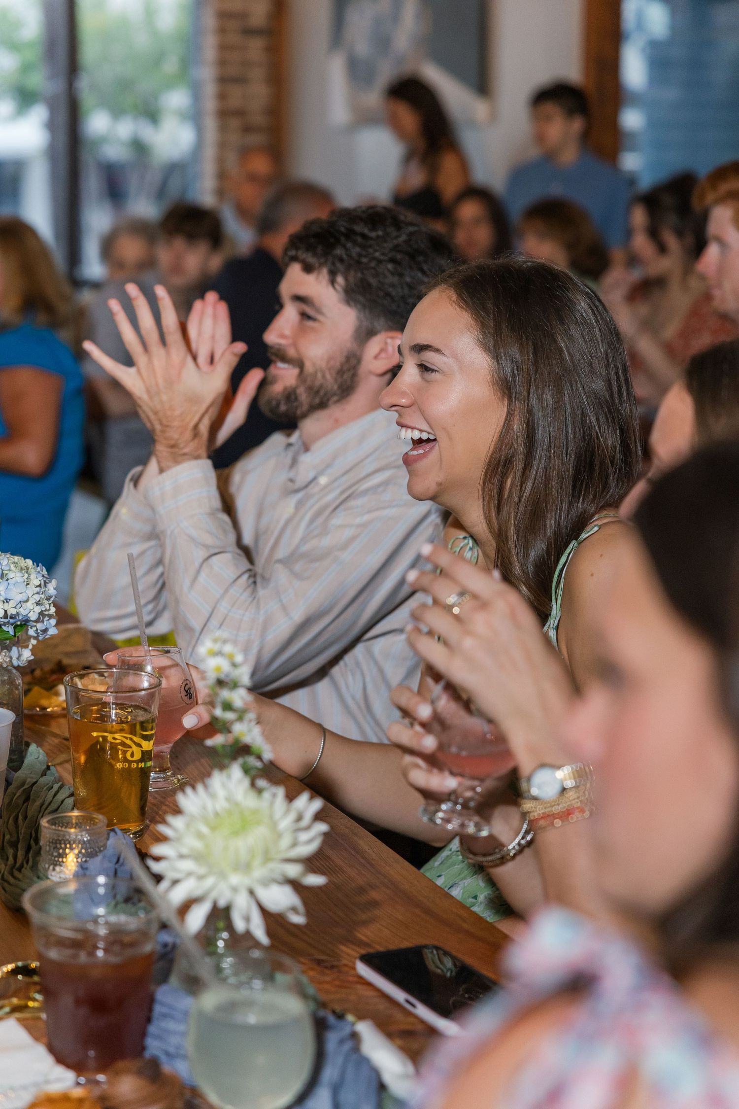 Guests laugh and clap joyfully at a lively indoor rehearsal dinner celebration.