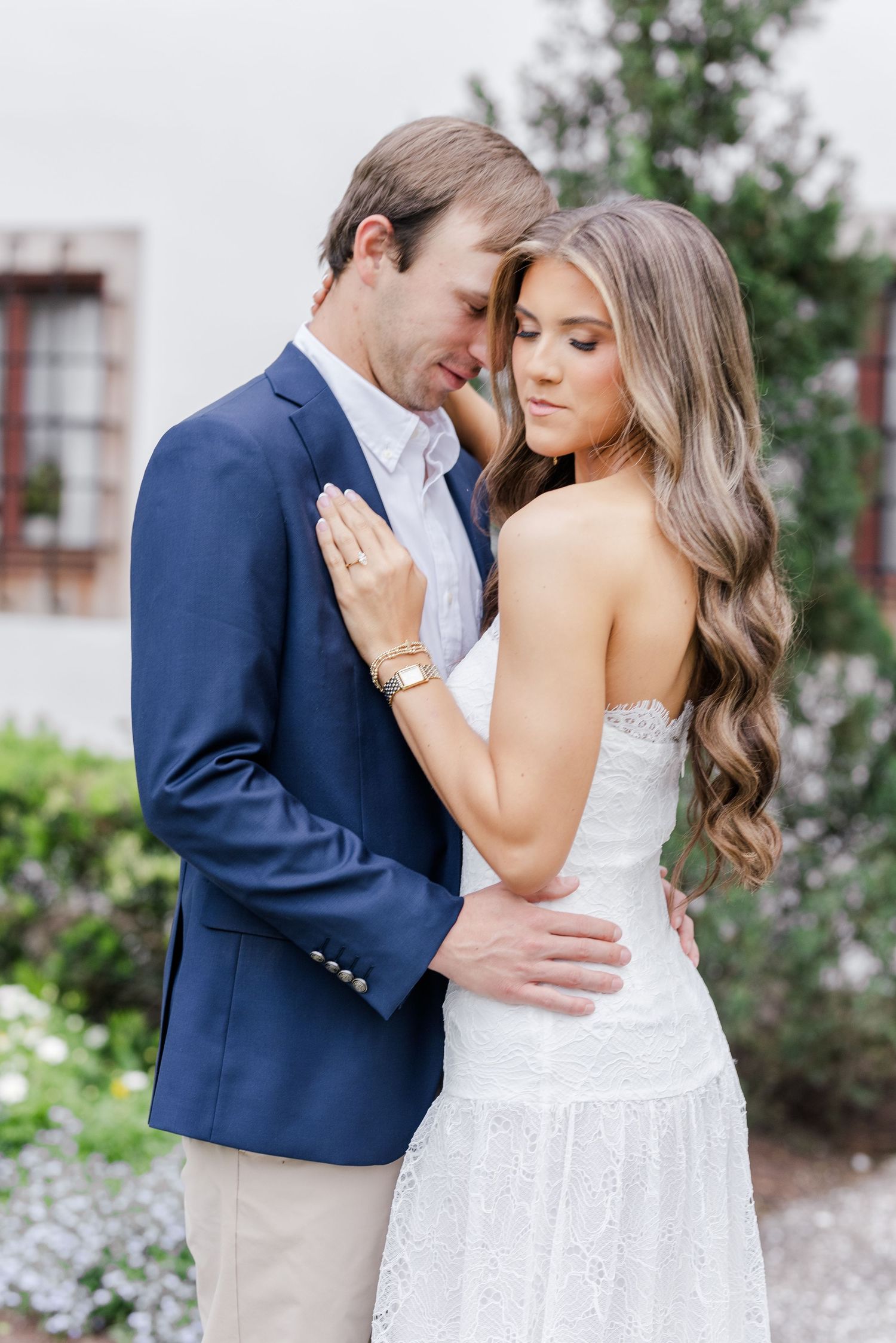 Engaged couple embracing outdoors, bride in white lace dress, groom in navy suit, garden backdrop.