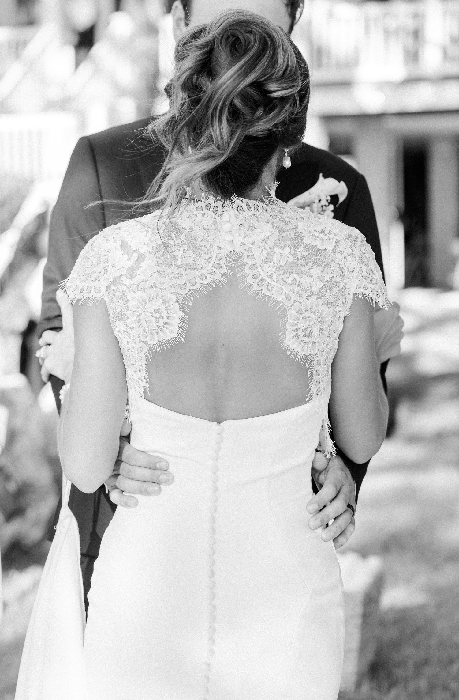 Black and white photo of bride in lace back wedding dress embraced by groom in suit at outdoor wedding.