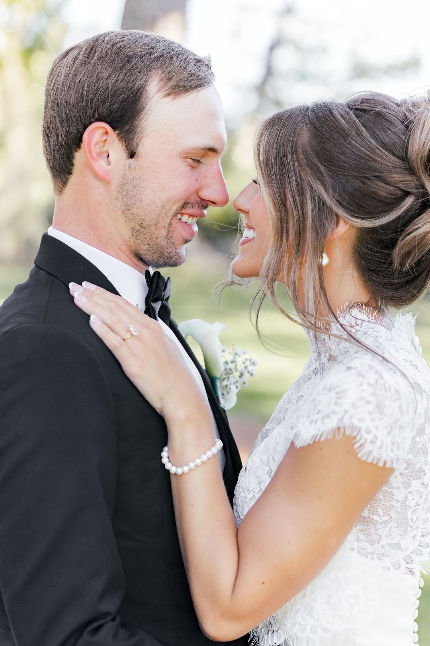 A smiling bride and groom in wedding attire share a romantic moment, with the bride wearing a lace dress and pearl bracelet.