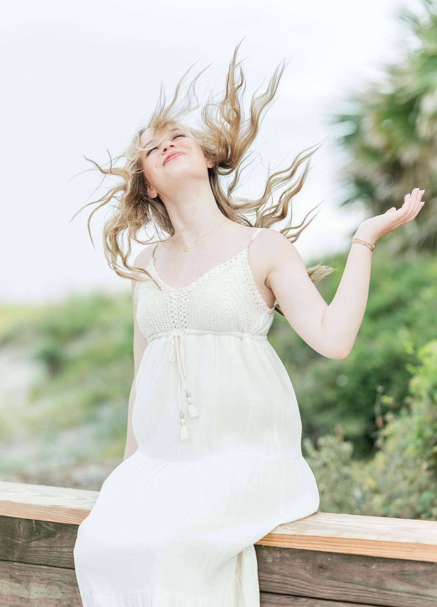 A figure in a flowing white dress with windswept hair poses joyfully against a bright sky in a carefree moment.