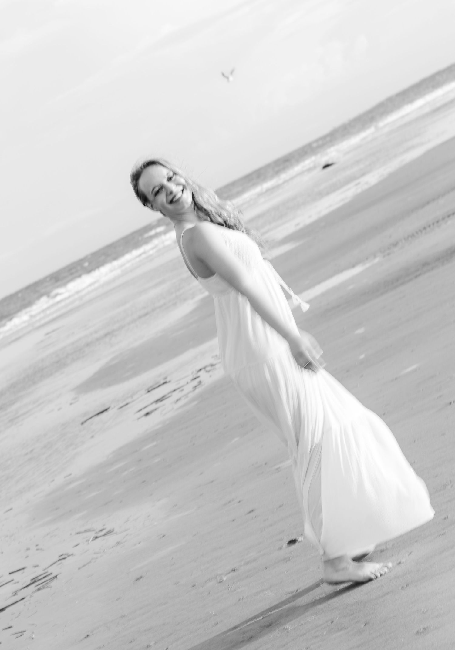 A joyful monochrome beach portrait shows movement in a flowing white dress against the sand.