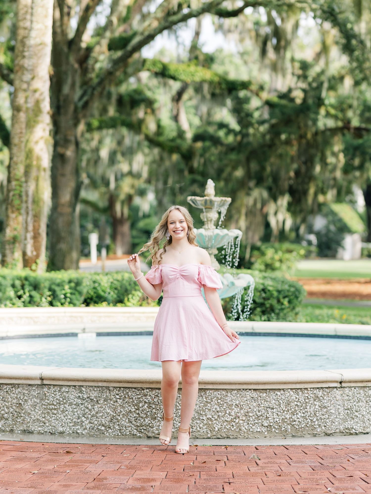 A pink off-shoulder dress is worn near an ornate fountain in a lush garden setting with Spanish moss-covered trees.