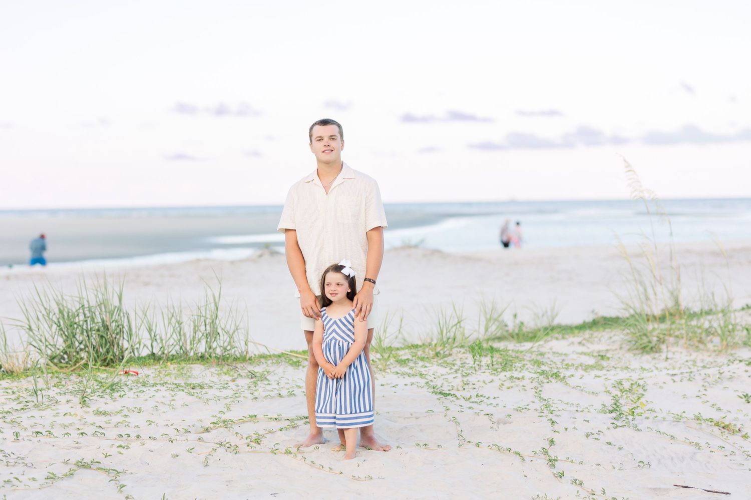 A parent and child stand together on a sandy beach dune with ocean views in the background on a cloudy day.