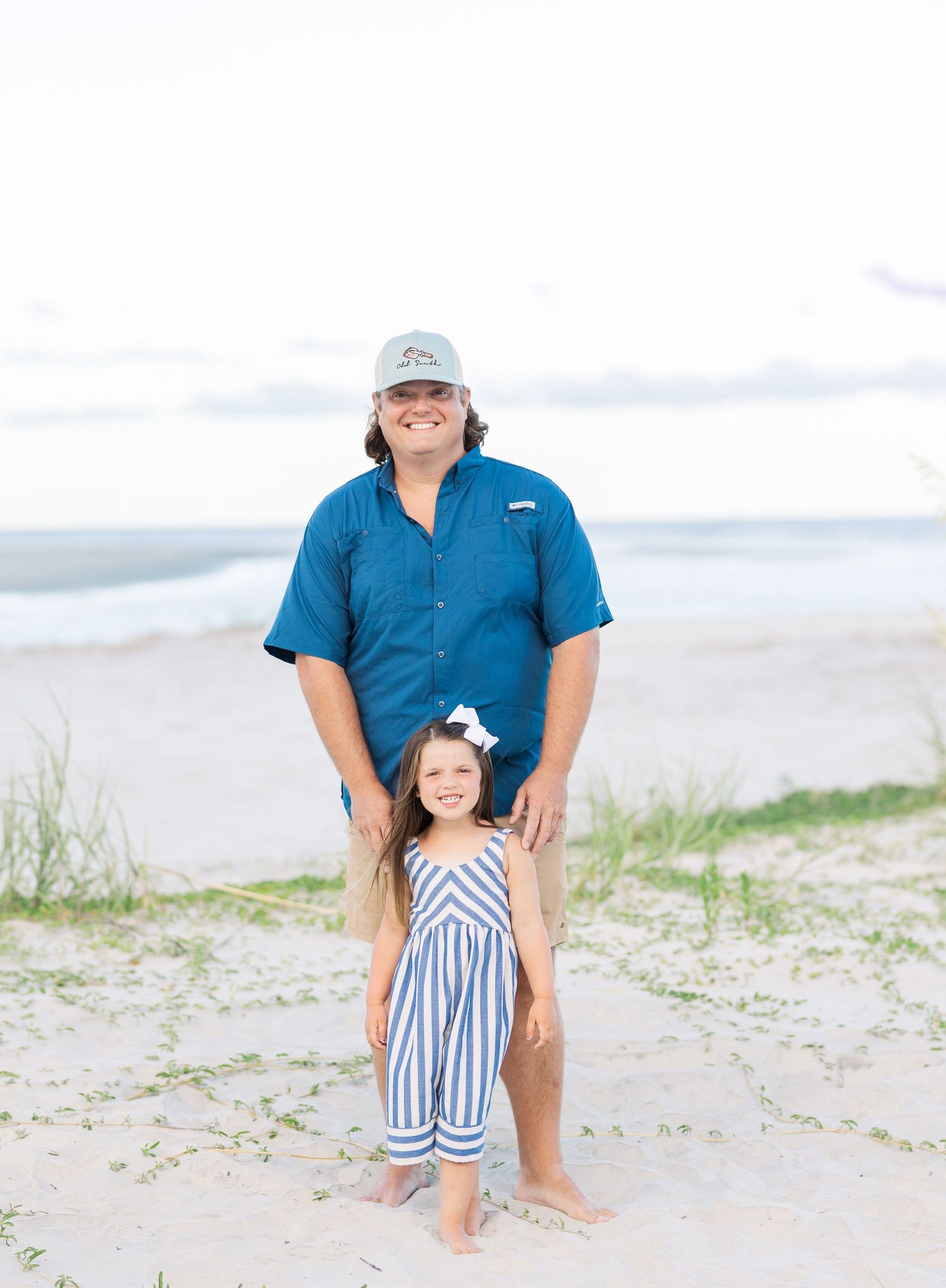 Two people standing together at a sandy beach with ocean waves and dunes in the background.