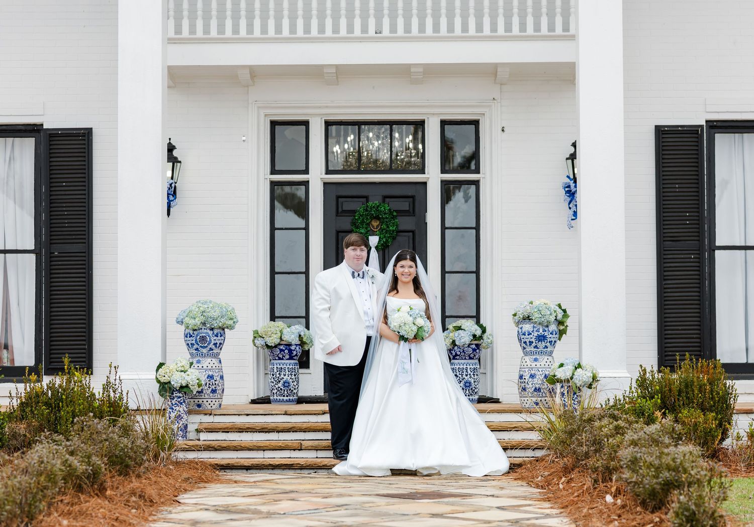 Bride and groom pose together in front of elegant white venue with blue and white floral arrangements.