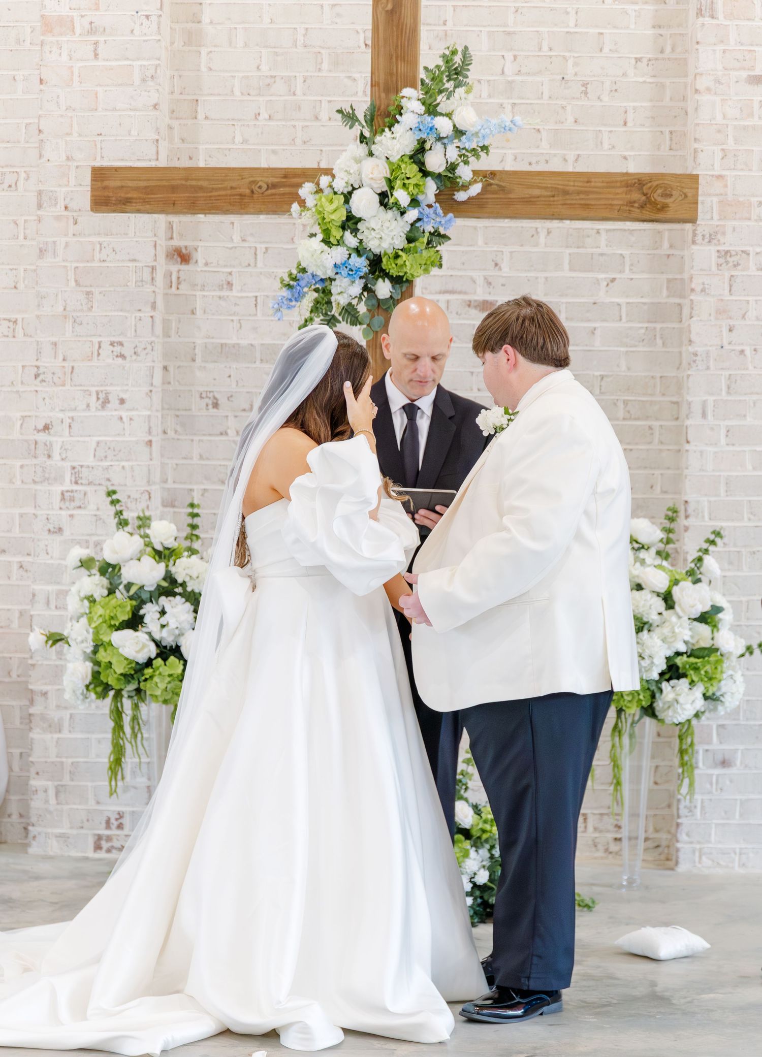 Couple holds hands during wedding ceremony at altar with wooden cross and elegant blue and white flowers.