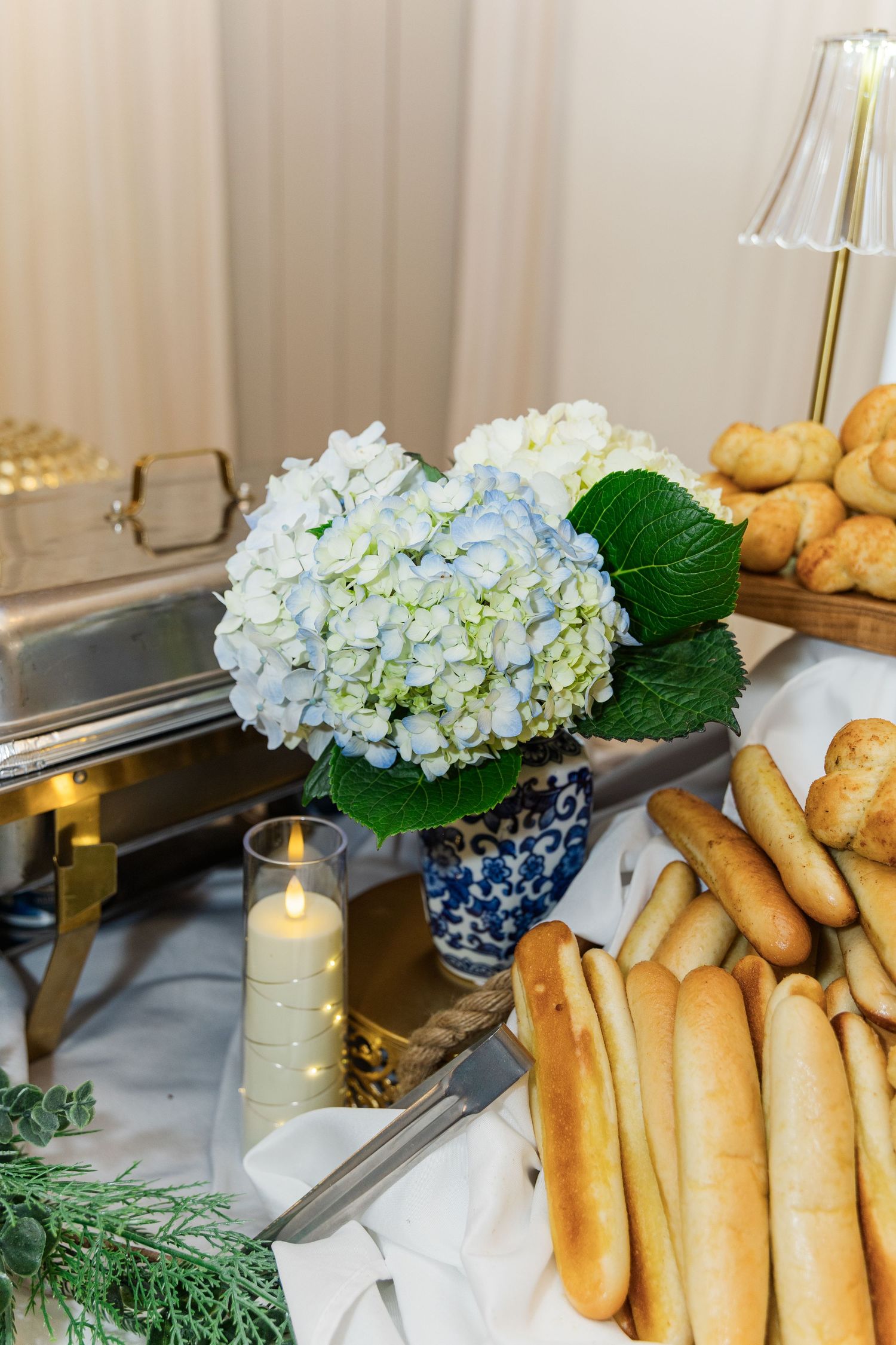 Blue and white hydrangea arrangement beside breadsticks and rolls on catering table.