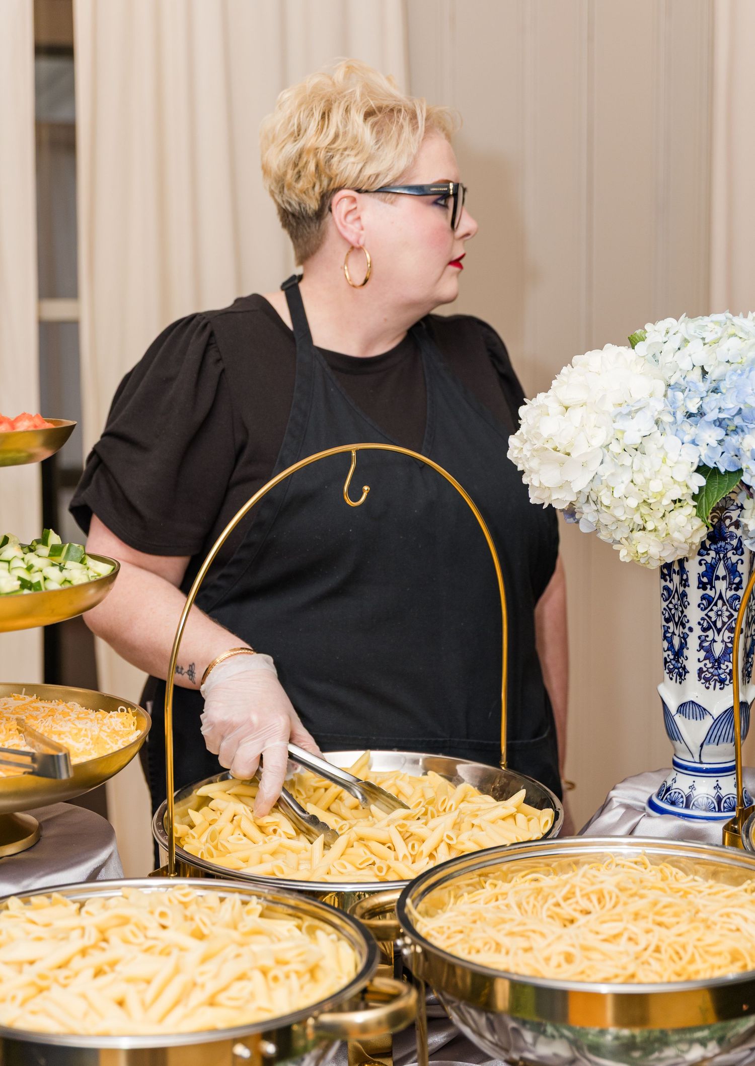 Catering staff member in black apron serves pasta dishes at wedding reception buffet with blue hydrangea decor.