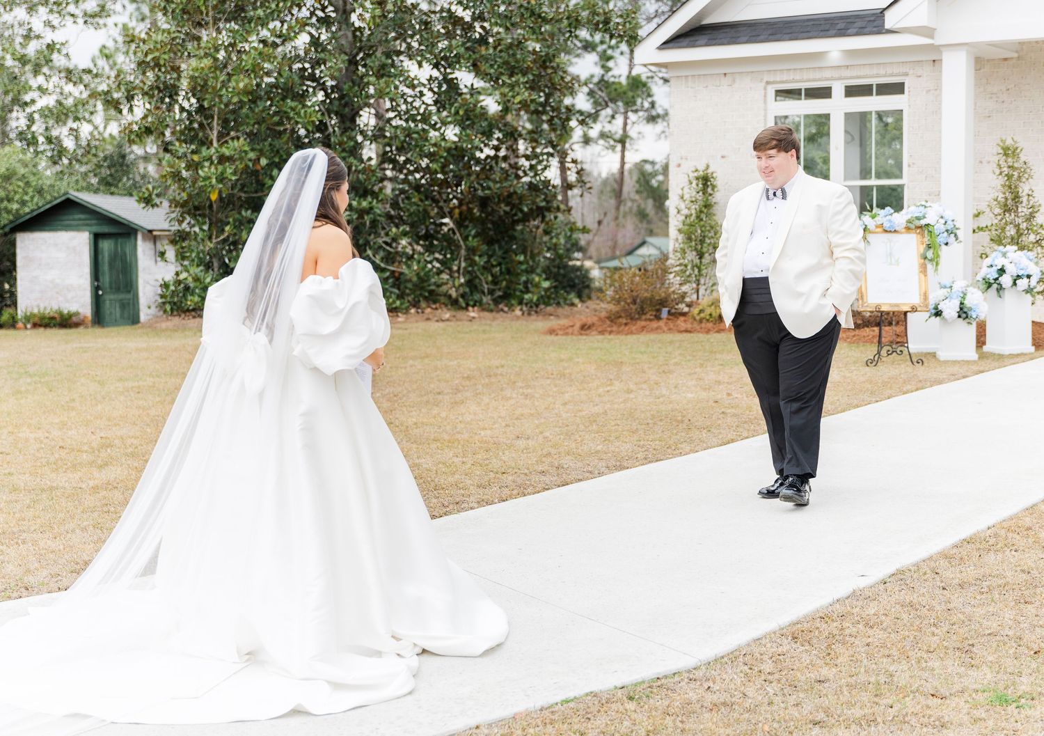 Groom in white tuxedo jacket turns for a first look at his bride in a flowing white gown on a garden pathway.