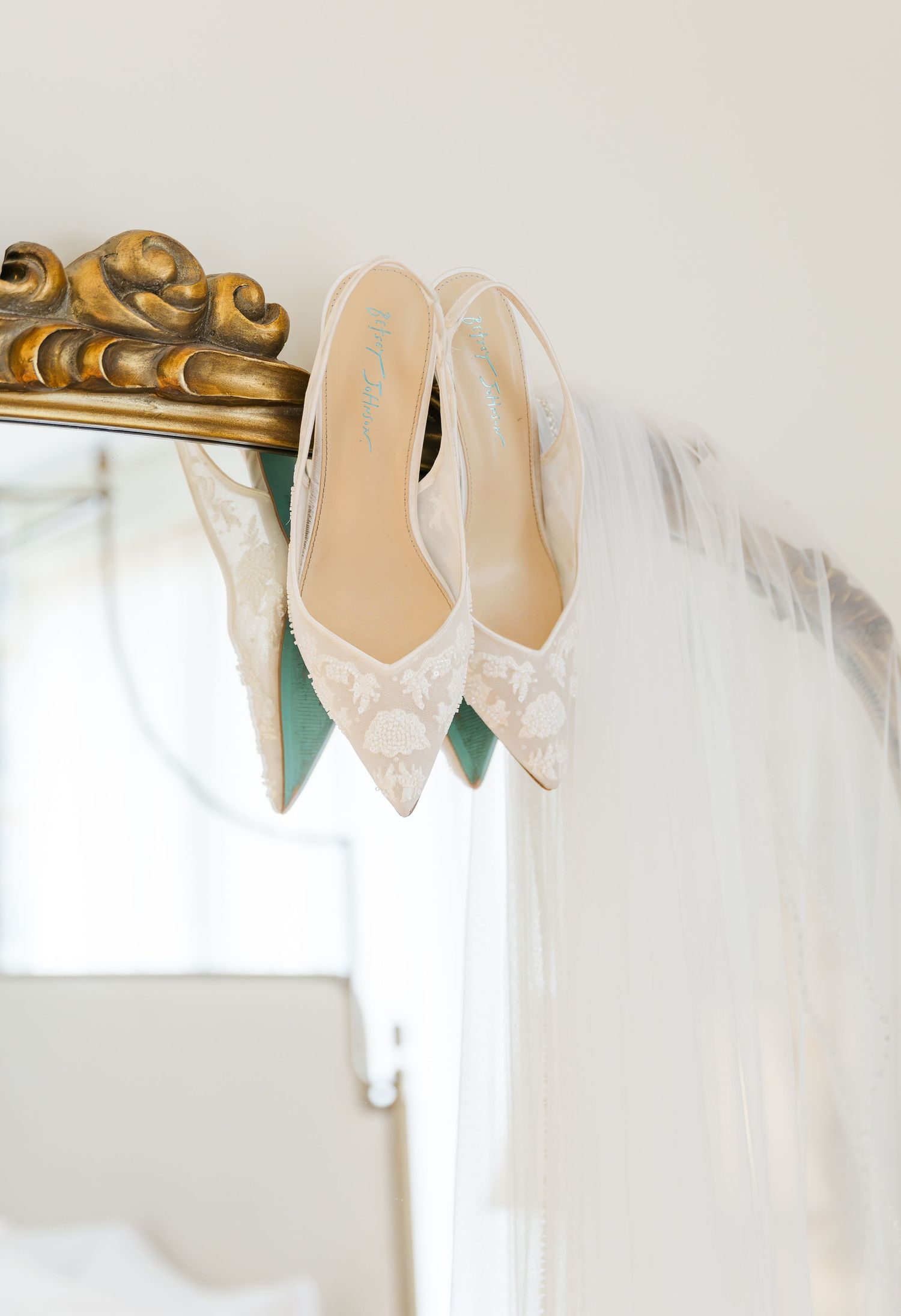 Lace bridal heels with teal soles hanging on gold ornate mirror with veil.