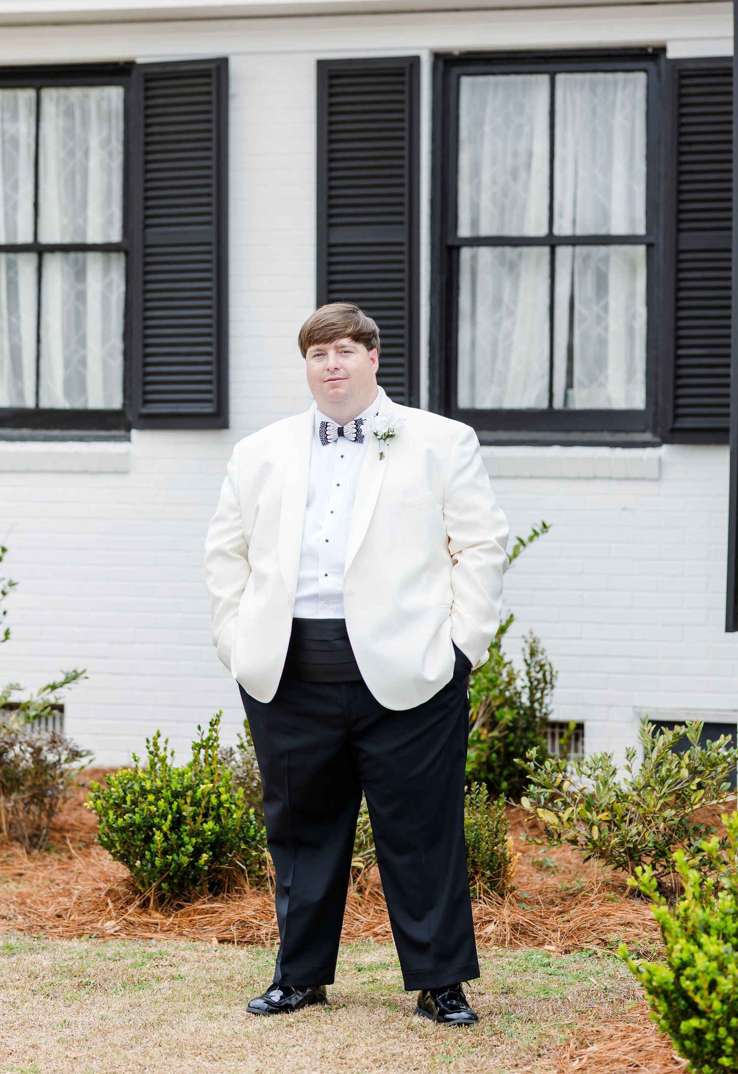 Groom in white tuxedo jacket and black bow tie standing outside a white house with black shutters.