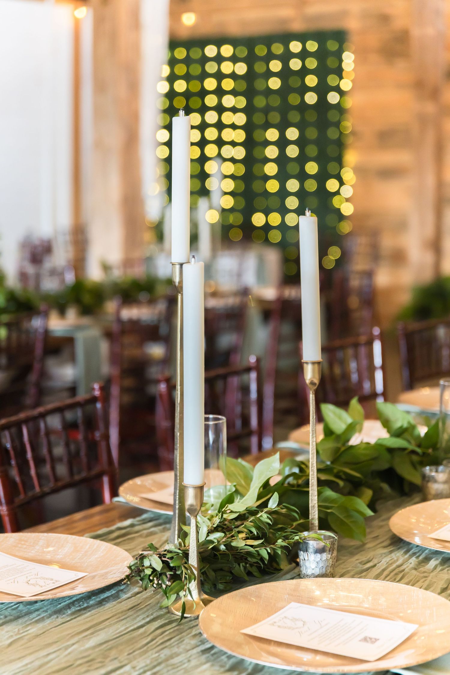Elegant wedding reception table setting with white candles, greenery, and place settings in warm lighting.