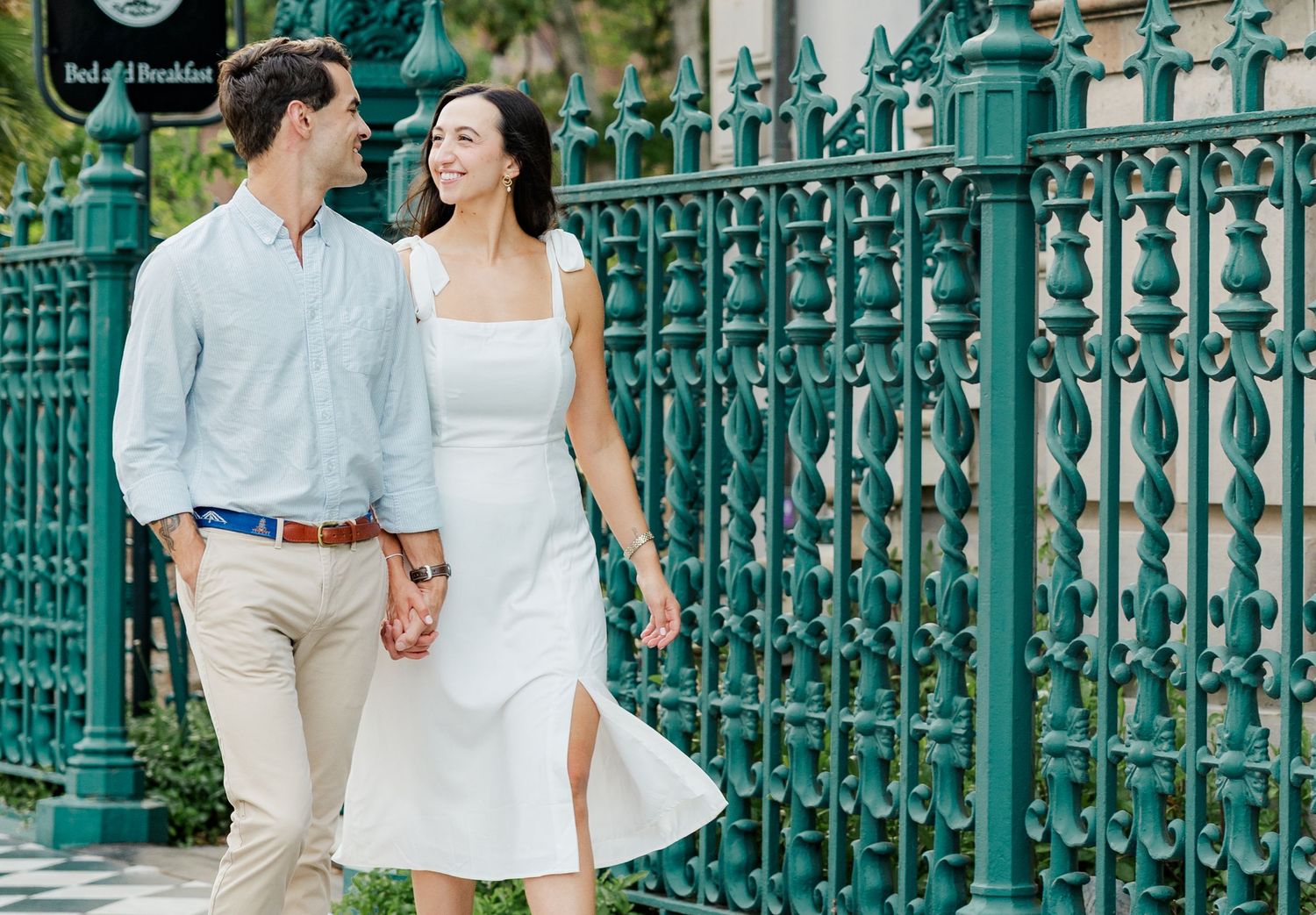 Couple holding hands and smiling while walking past ornate green iron fence at a bed and breakfast.